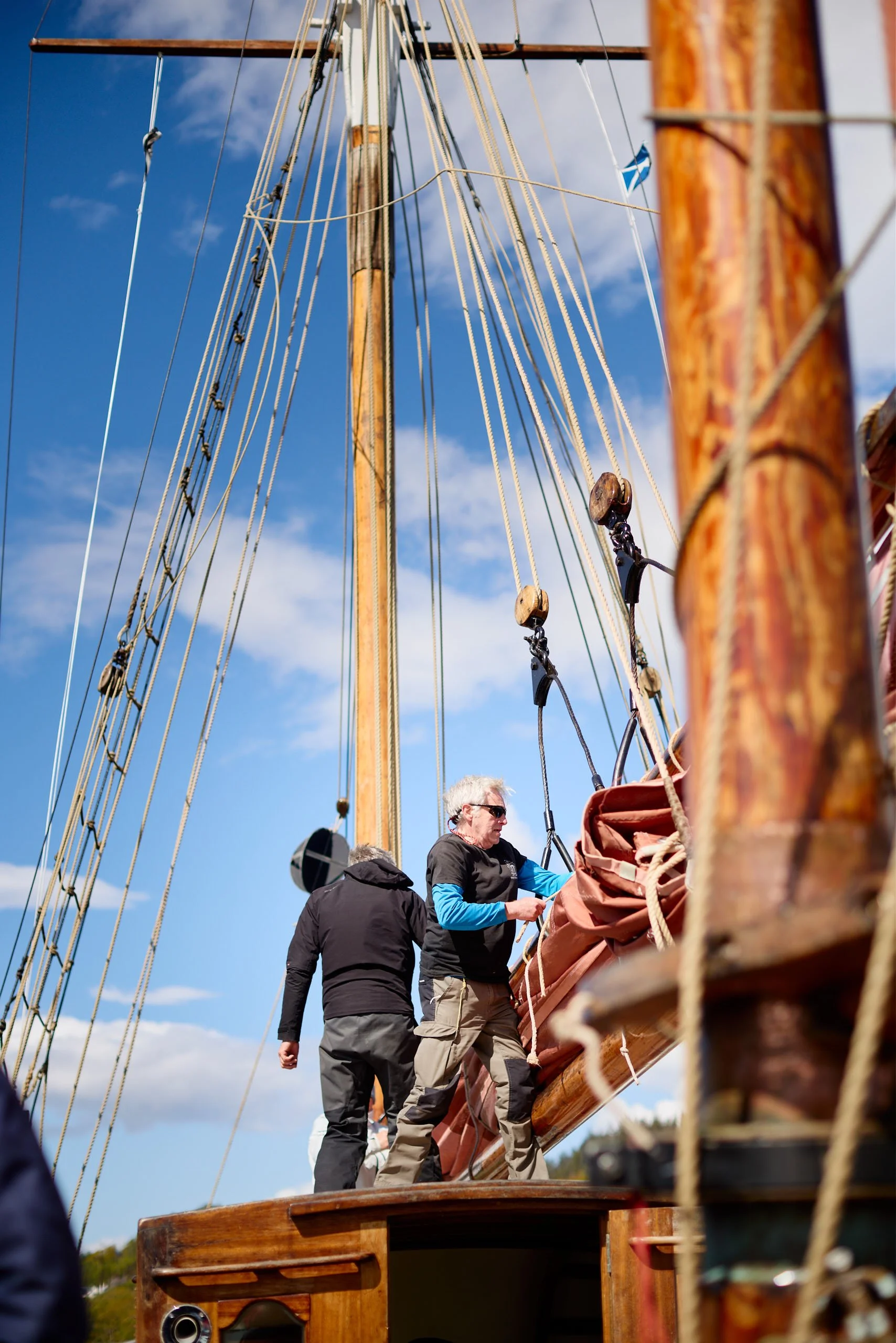 People on the deck of a wooden sailing ship, standing near the mast and rigging, with a partly cloudy sky in the background.