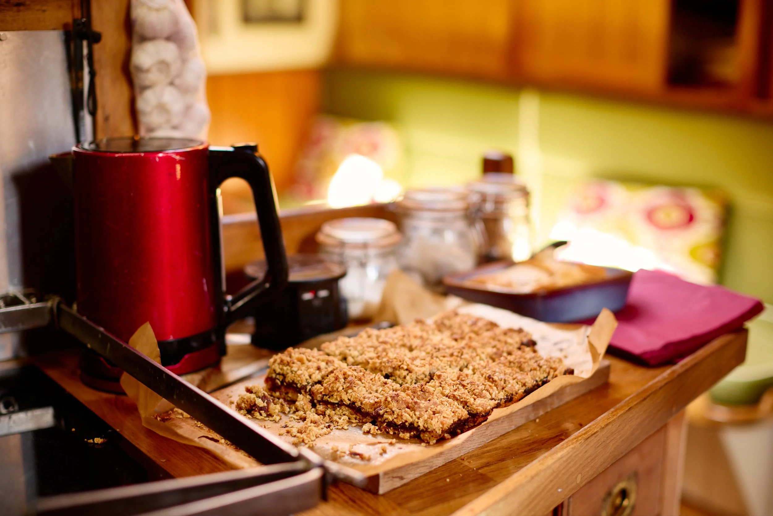 A tray with freshly baked crumb-topped bars on parchment paper, alongside a red coffee maker and jars of baking ingredients on a wooden countertop in a cozy kitchen.