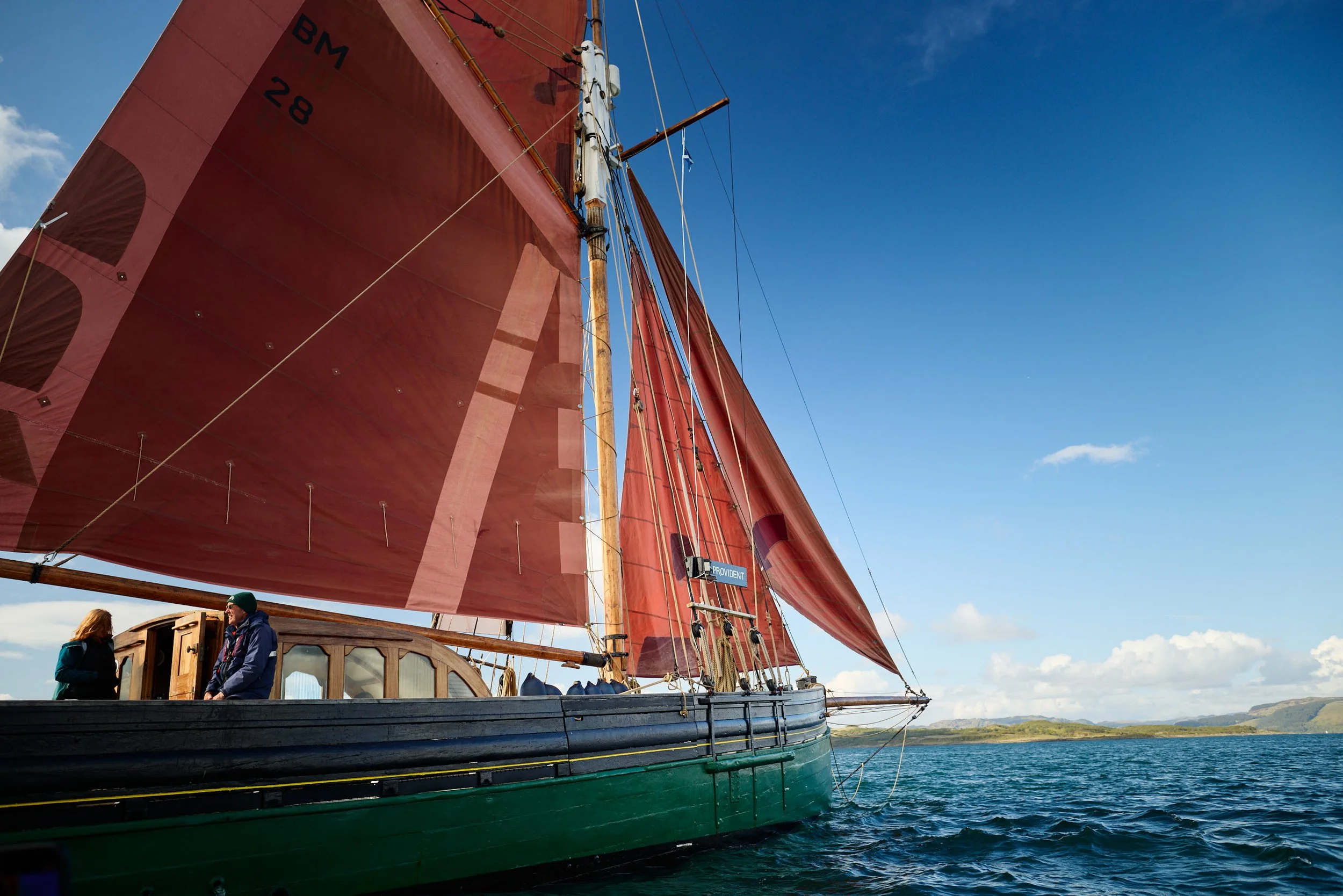 A large sailing ship with red sails on the water, with two people standing on the deck and a clear blue sky in the background.
