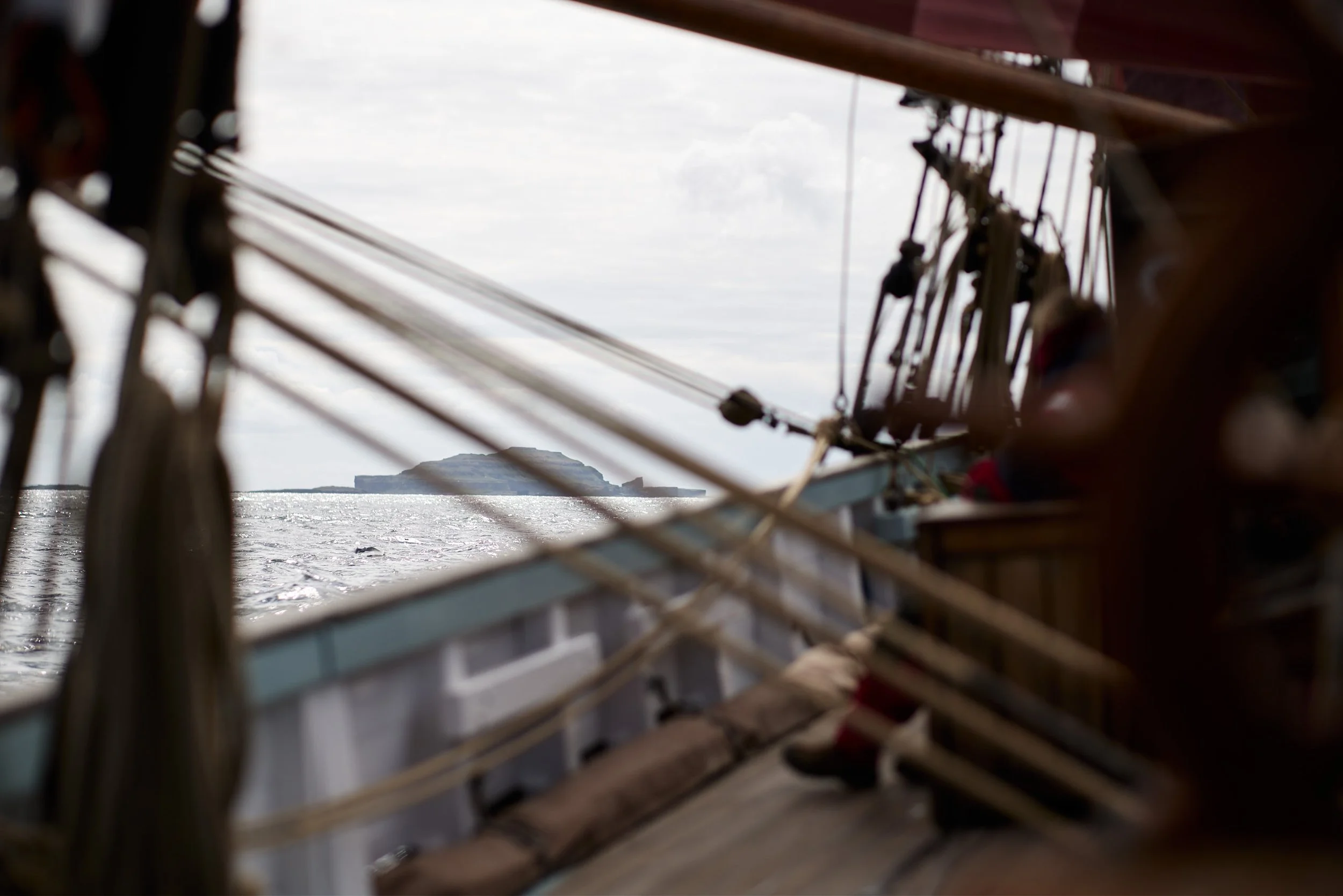View through the gap in a boat's wooden side, showing water and distant landmass with a cliff or hill in the background on a cloudy day.