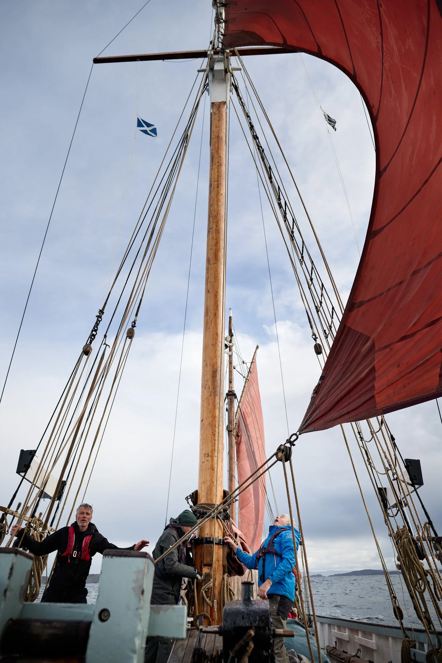 People working on the deck of Provident with tall masts and red sails, facing the sea with a cloudy sky.