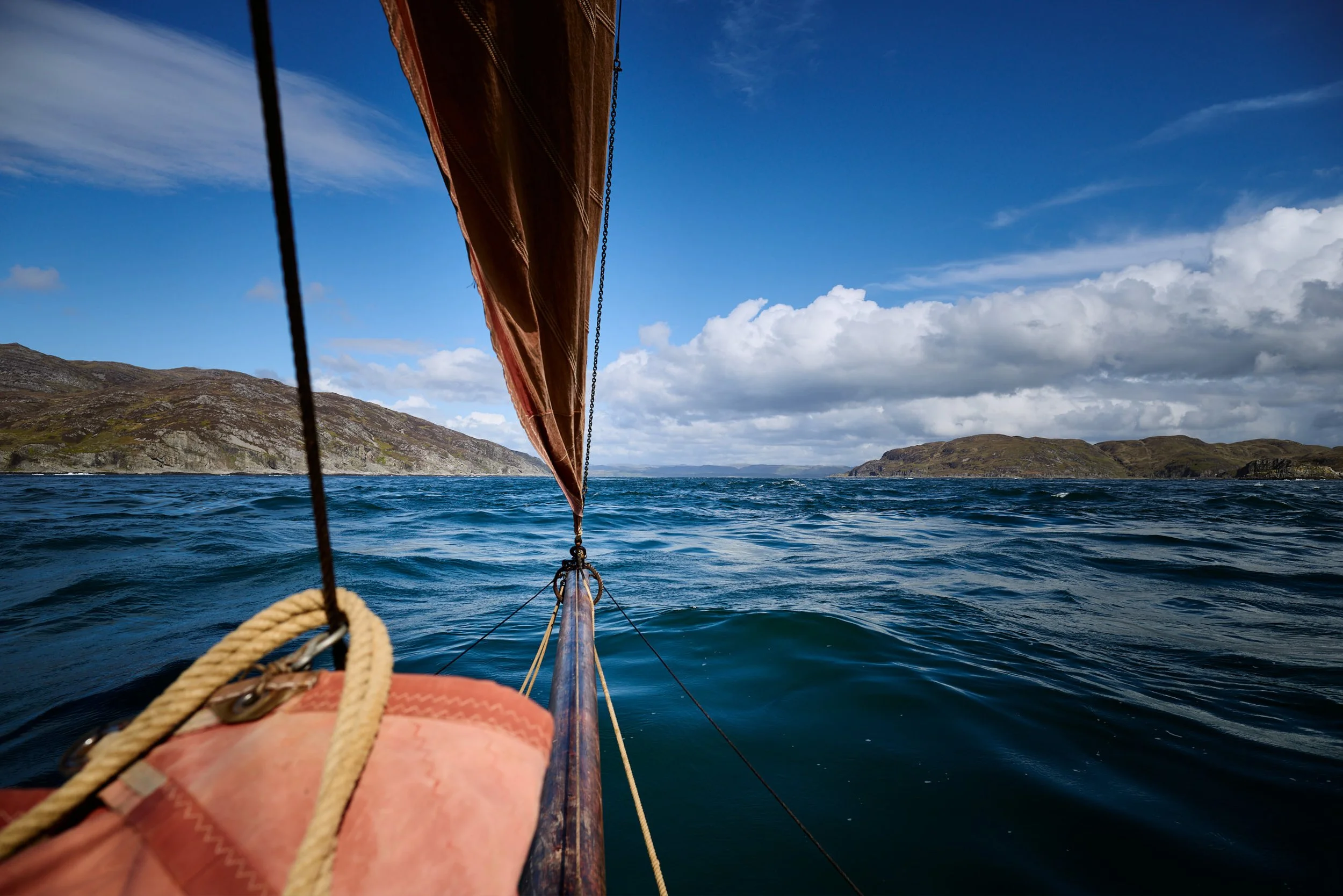 View from the bow of a boat sailing on open water with distant landforms and a mostly blue sky with some white clouds.