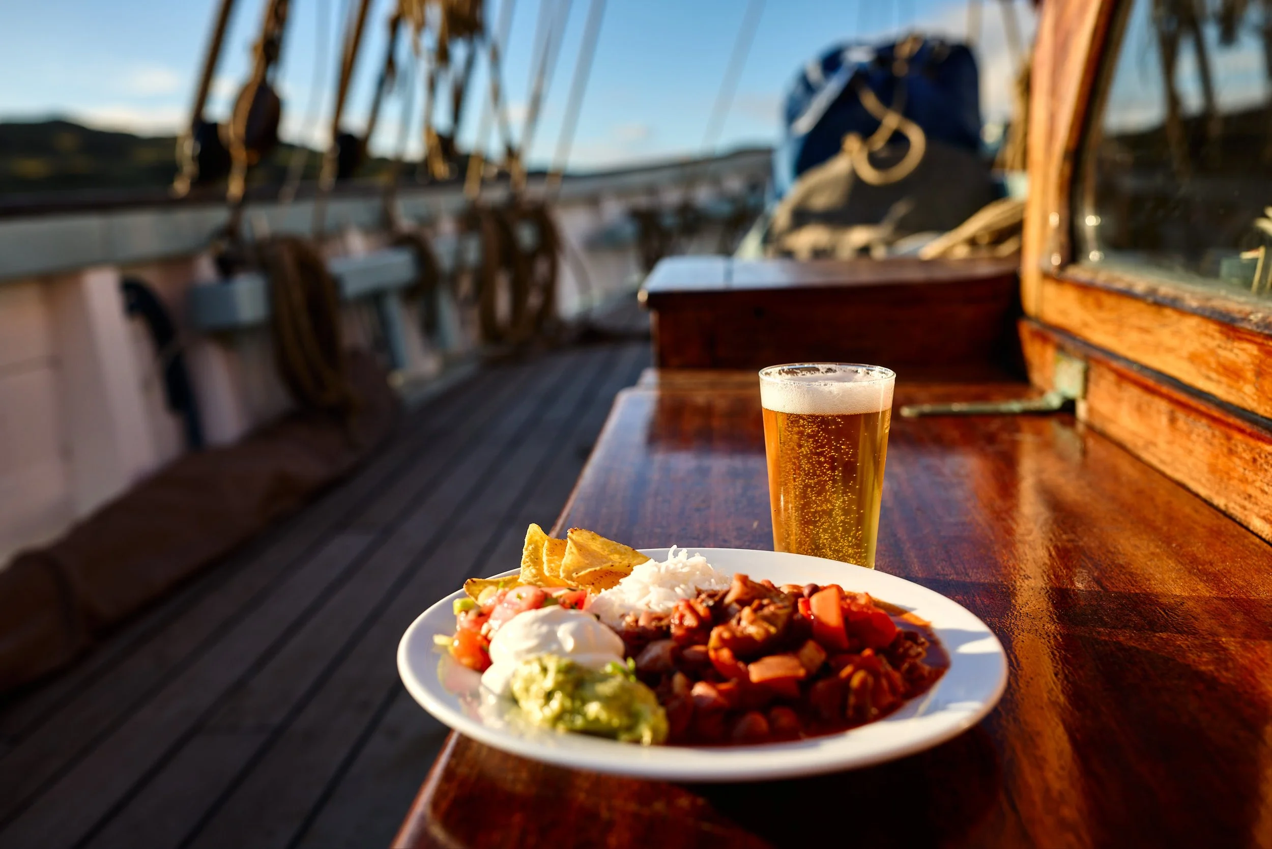 A plate of Mexican food, a glass of beer, and a slice of lime on a wooden table aboard a boat during sunset.