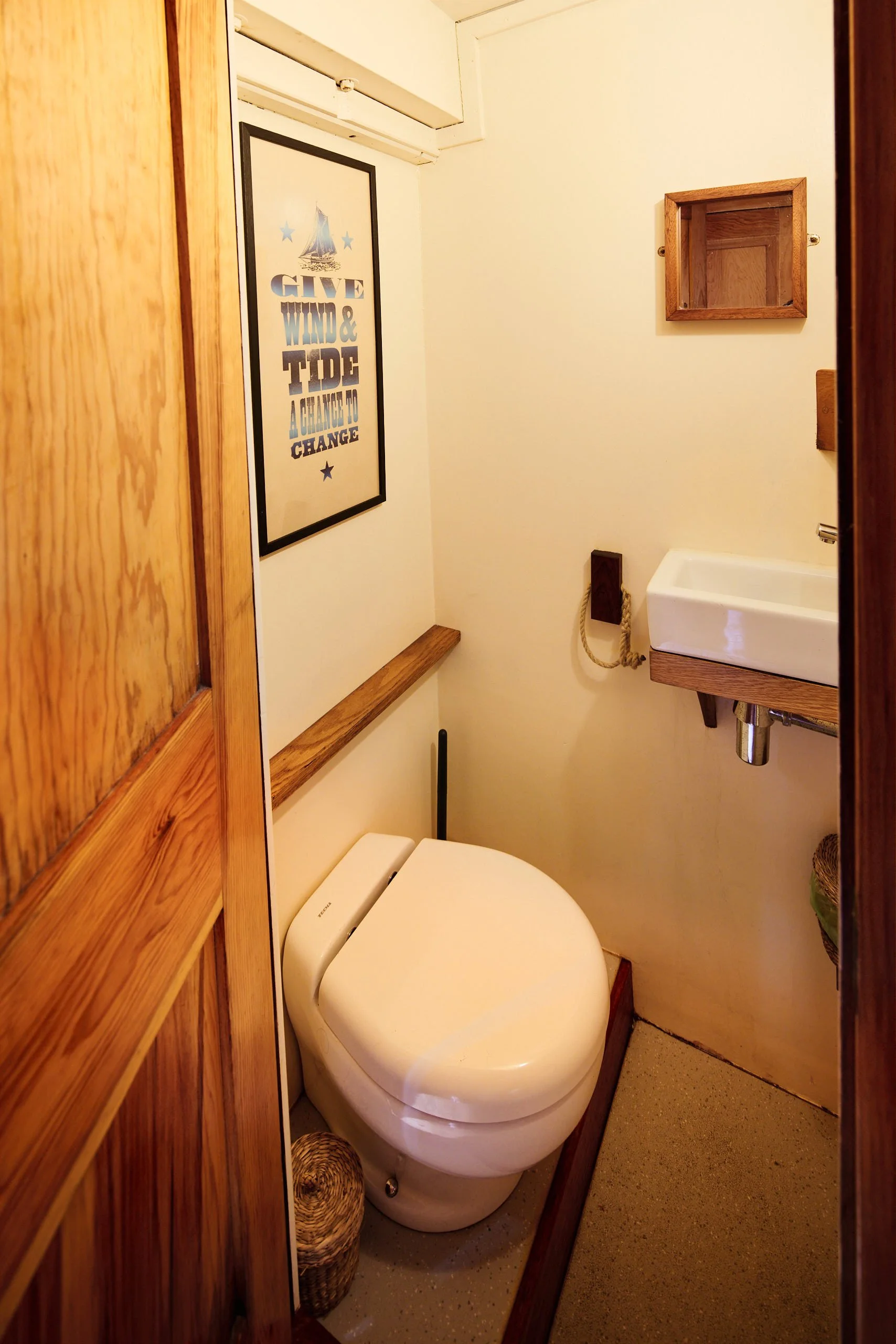 Small bathroom with a modern white toilet, wooden accents, wall art with a sailboat and quote, small sink, wooden shelf, and a framed empty box on the wall.