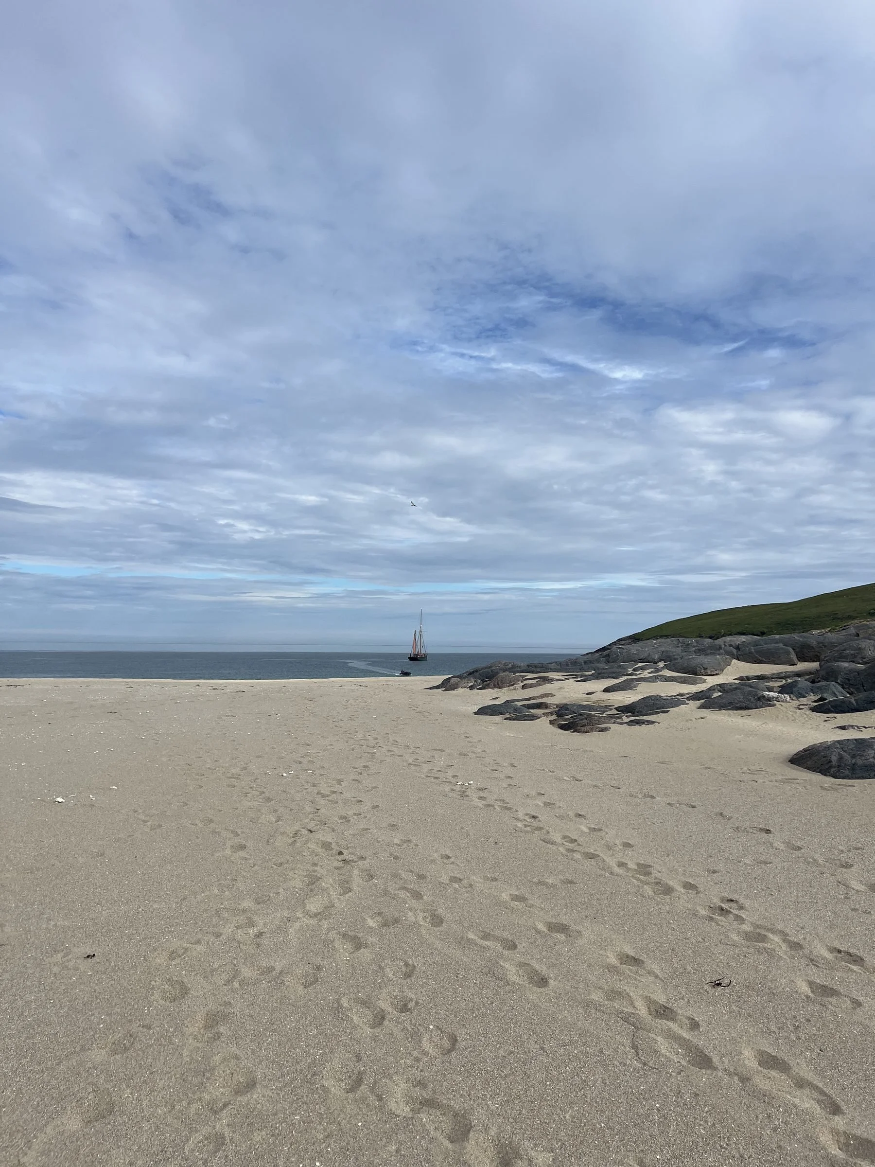 A sandy beach with multiple footprints, rocks on the right, a grassy hill in the background, and a sailboat on the ocean under a cloudy sky.