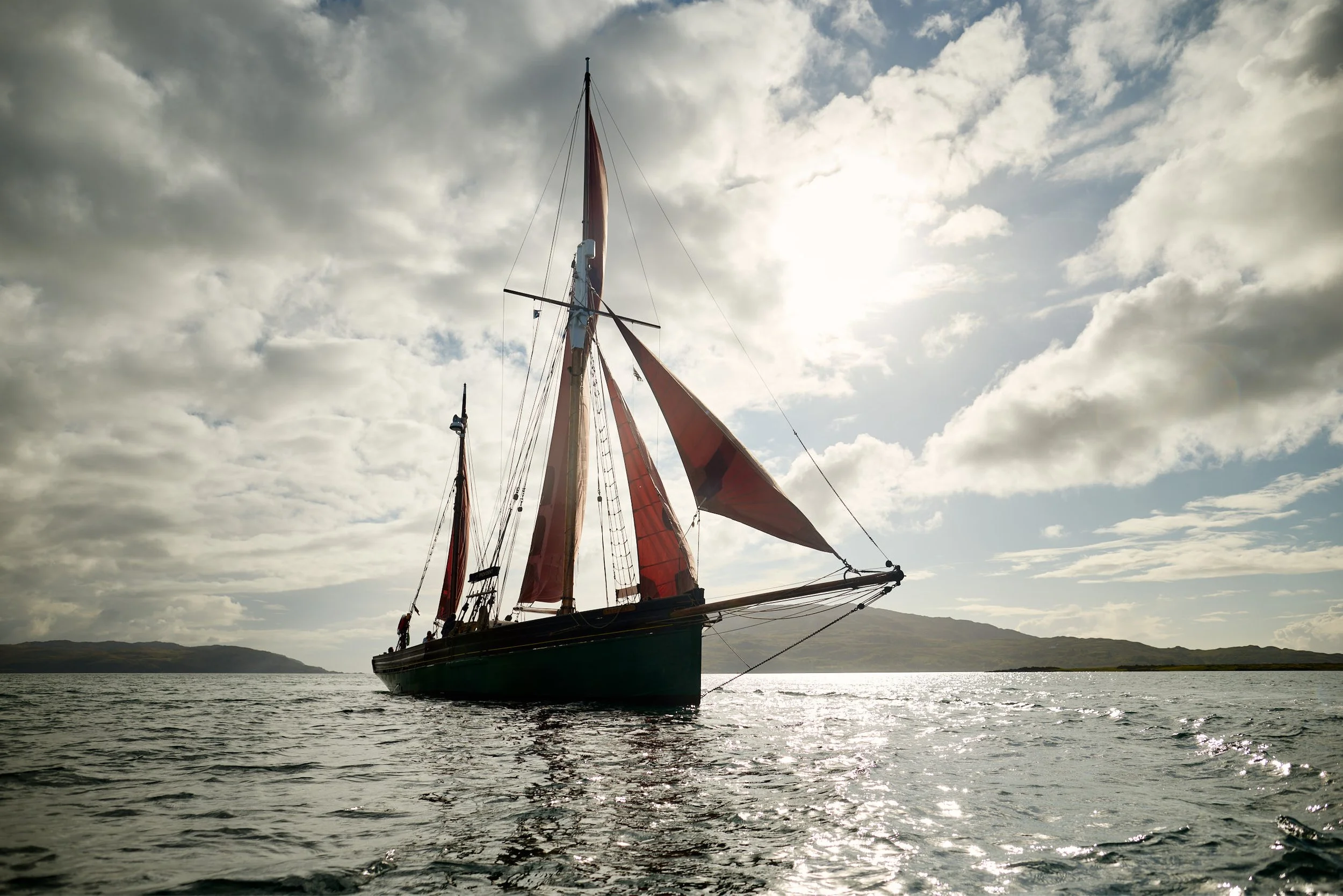 The sailboat, Provident,  with red sails floating on a body of water under a cloudy sky, with mountains in the distance.