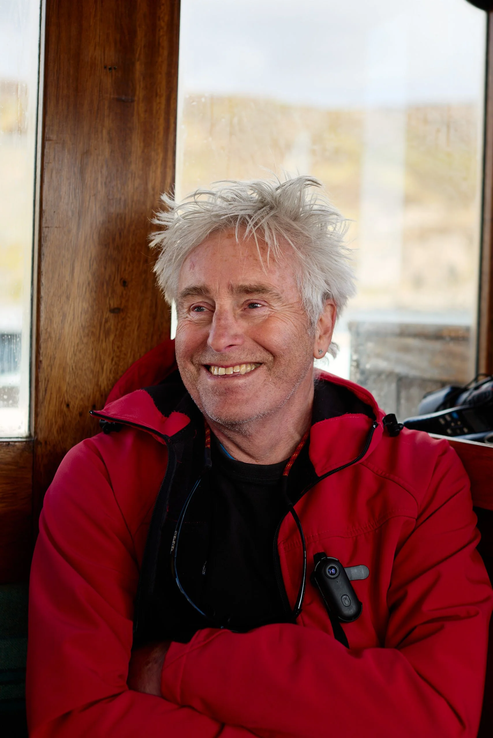 A man with white, messy hair smiling, wearing a red jacket, sitting in front of a window with wooden framing, with a blurred outdoor landscape in the background.