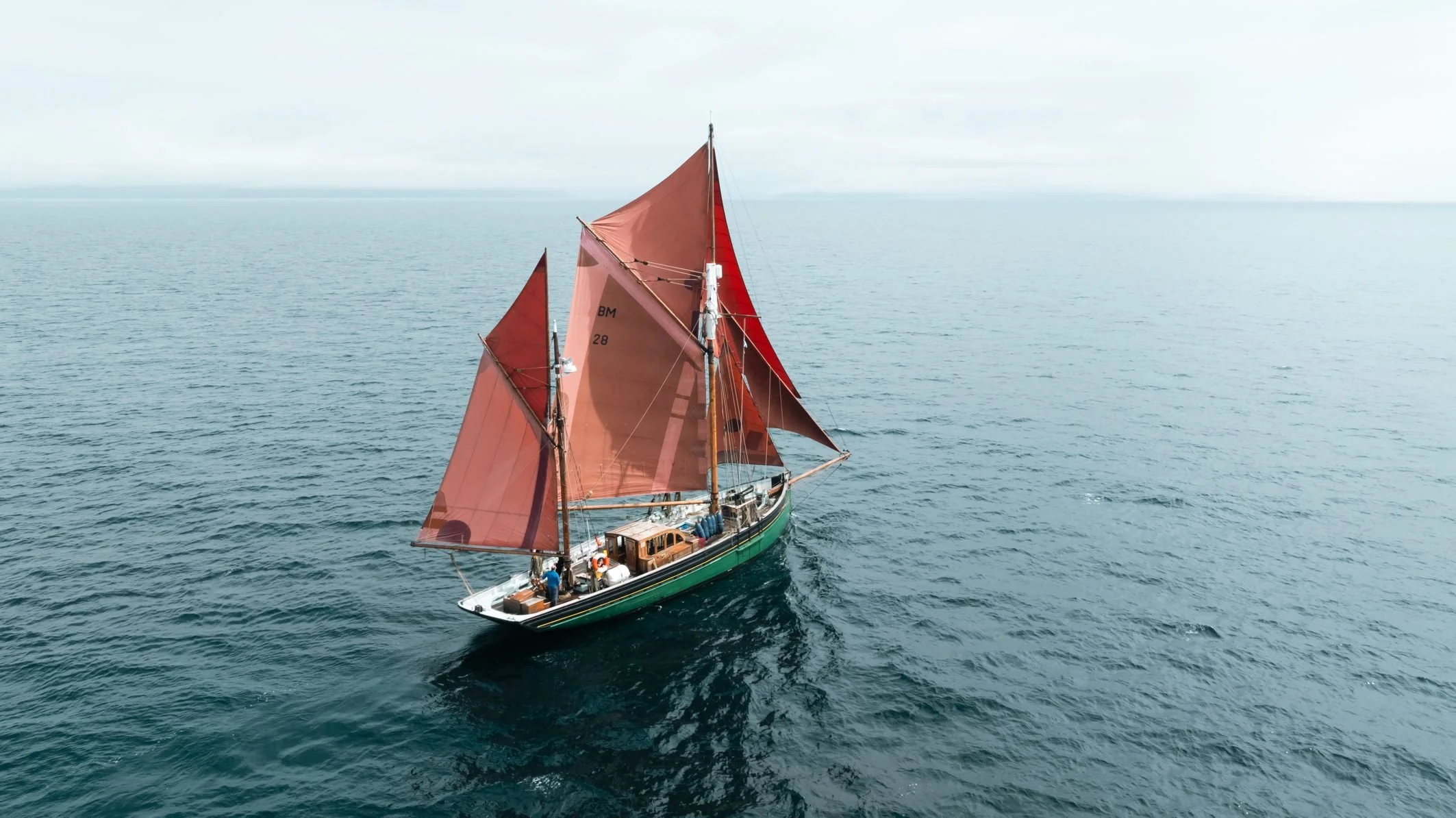 A sailboat with large reddish sails on calm open water under a cloudy sky.