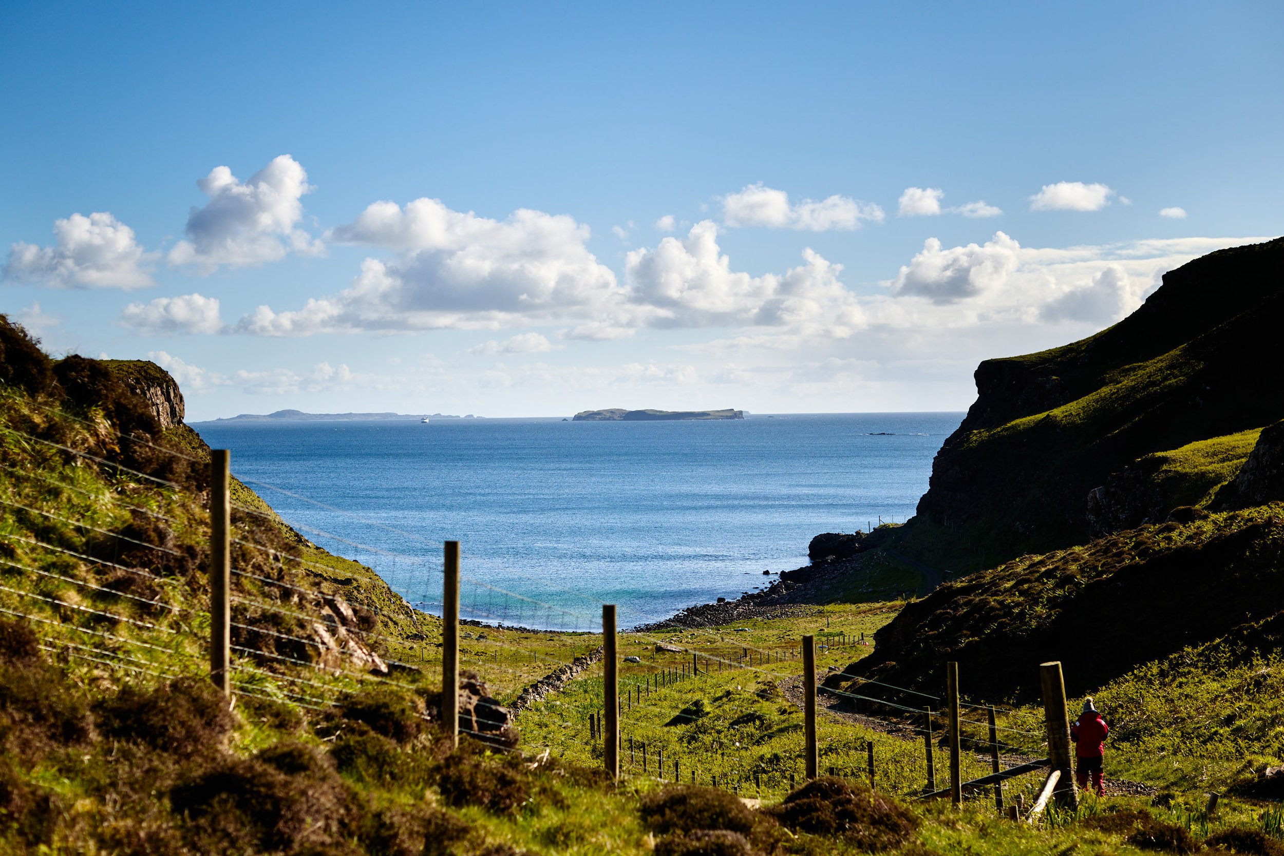 A coastal landscape with grassy hills on either side, a blue ocean with small islands on the horizon, partly cloudy sky, and a person walking along a trail in the foreground.
