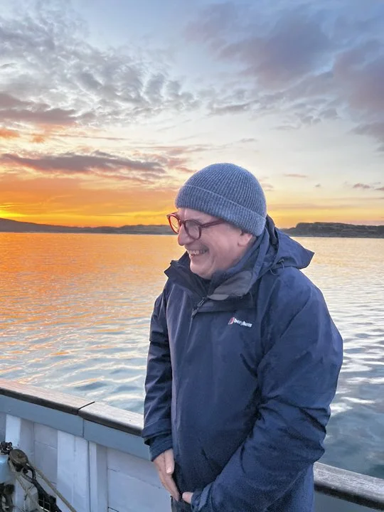 A man wearing glasses, a gray beanie, and a navy jacket standing on a boat at sunset, with a body of water and a colorful sky in the background.