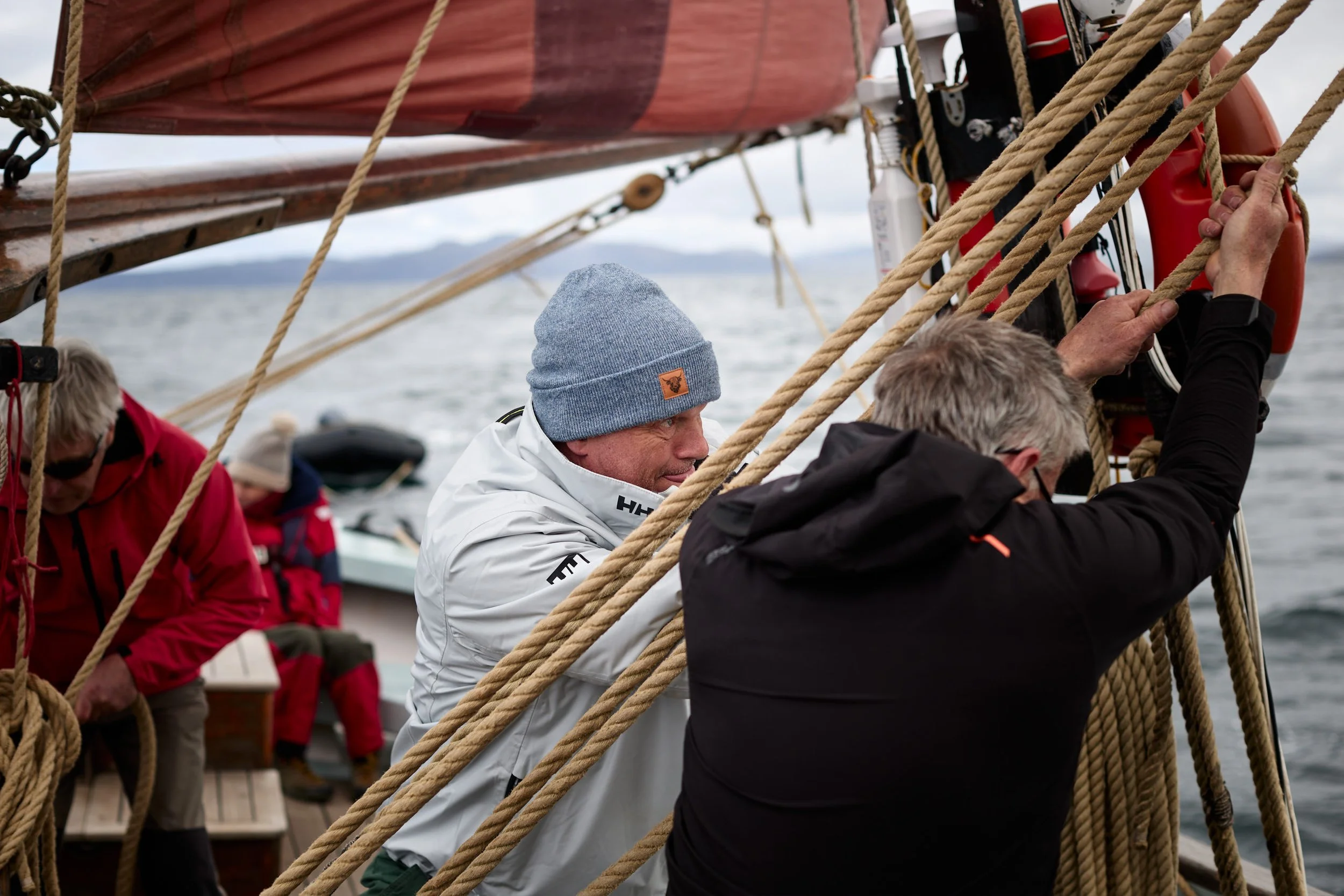 People working together on a sailboat, handling ropes and equipment, with a view of water and mountains in the background.