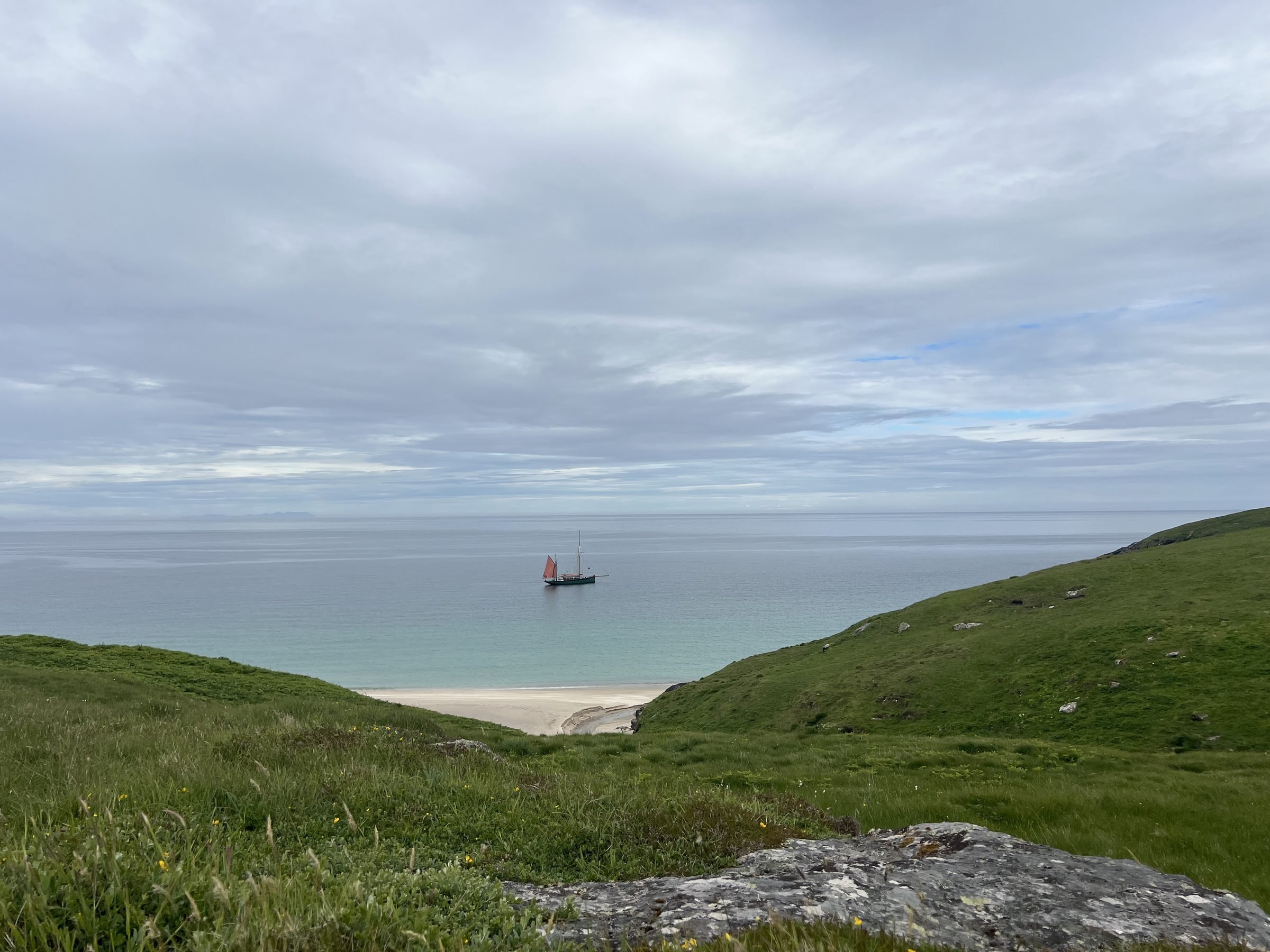The sailboat, Provident,  with a red sail moving across calm waters under a cloudy sky, with green hills in the foreground on Barra beach.