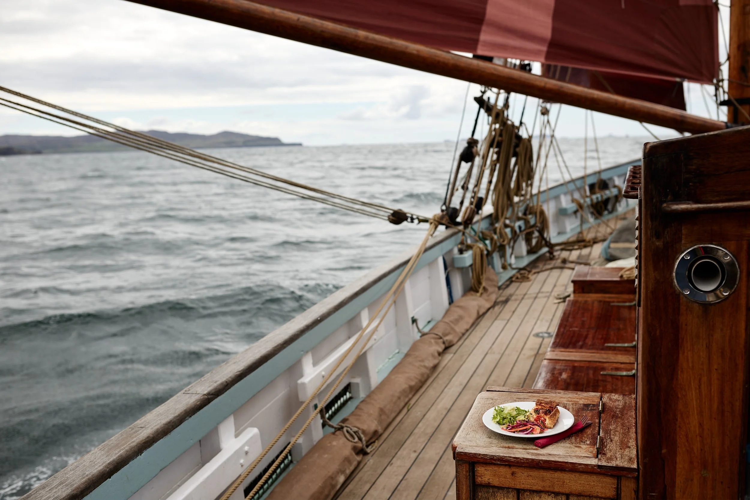 View from a wooden boat looking out at the ocean with land in the distance. There is a small table with a plate of food, including a slice of pie, and green salad.