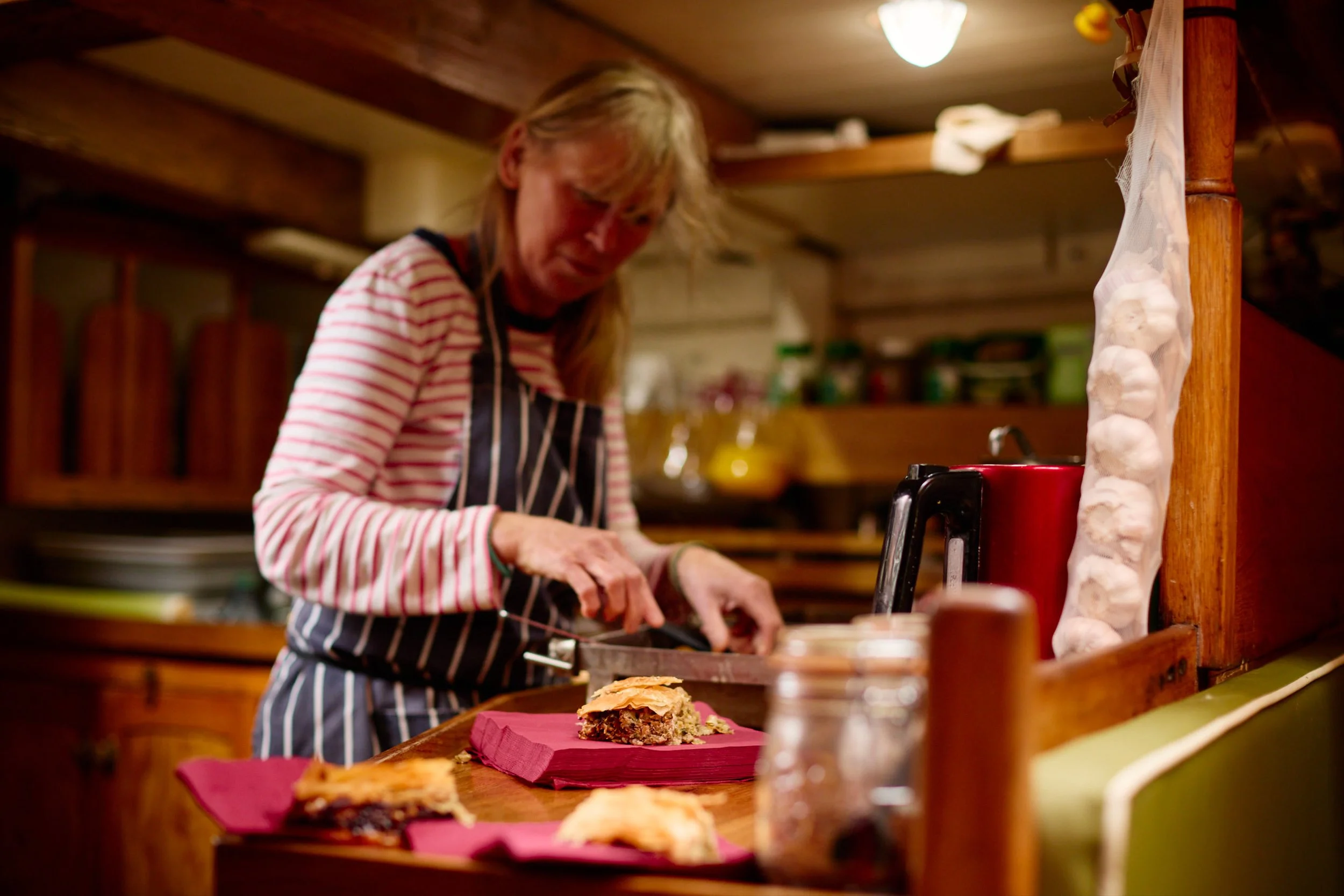 Woman in striped shirt and apron preparing food in a cozy kitchen with wooden cabinets and warm lighting.