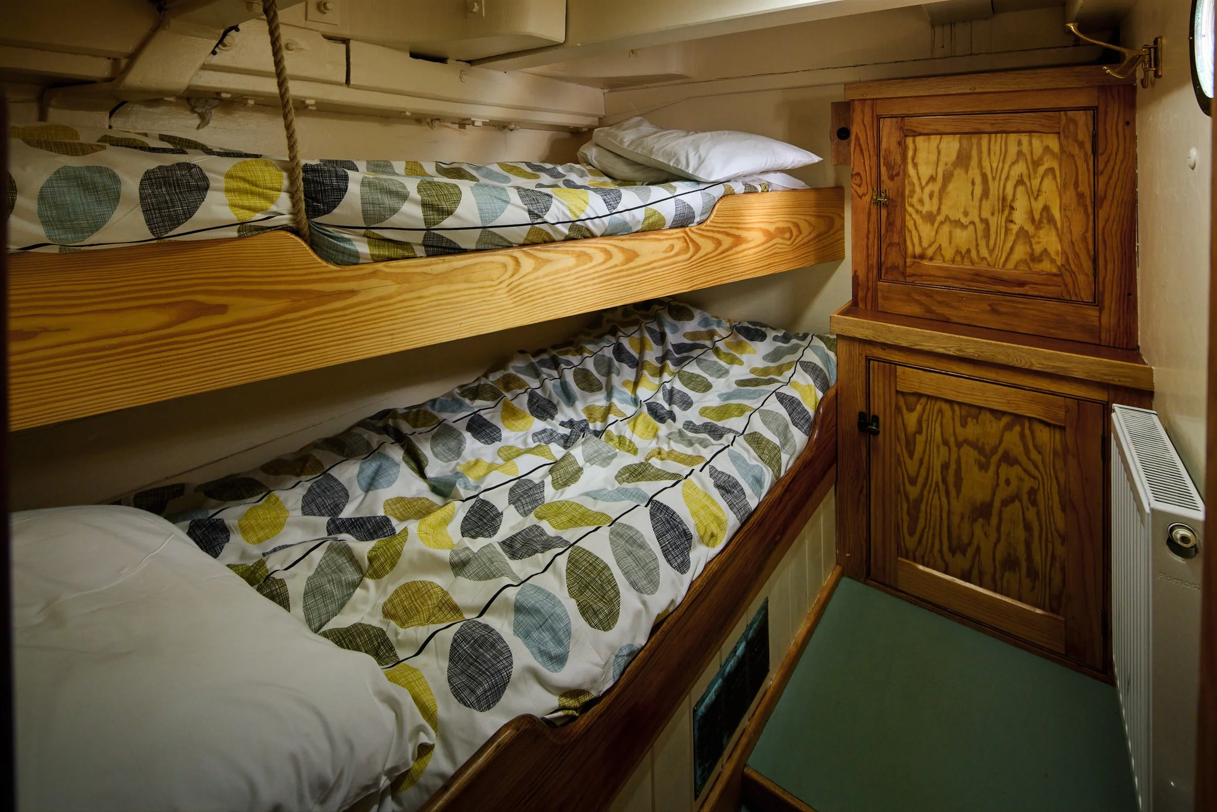 A narrow bunk bed with two beds, covered with sheets featuring a leaf pattern in shades of yellow, blue, black, and gray. There is a white pillow on the top bunk. To the right, there is a wooden cabinet with a door and a small latch, beneath a pole w