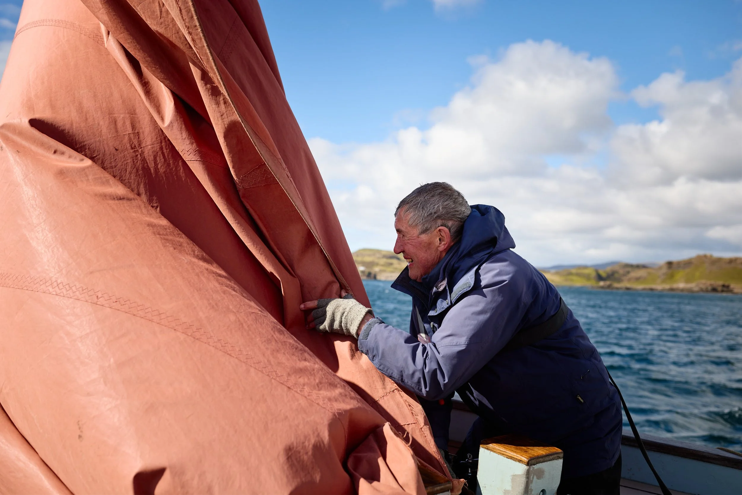 A man wearing a jacket and gloves is working on a large, orange boat or marine vessel outdoors near water, with a landscape and cloudy sky in the background.