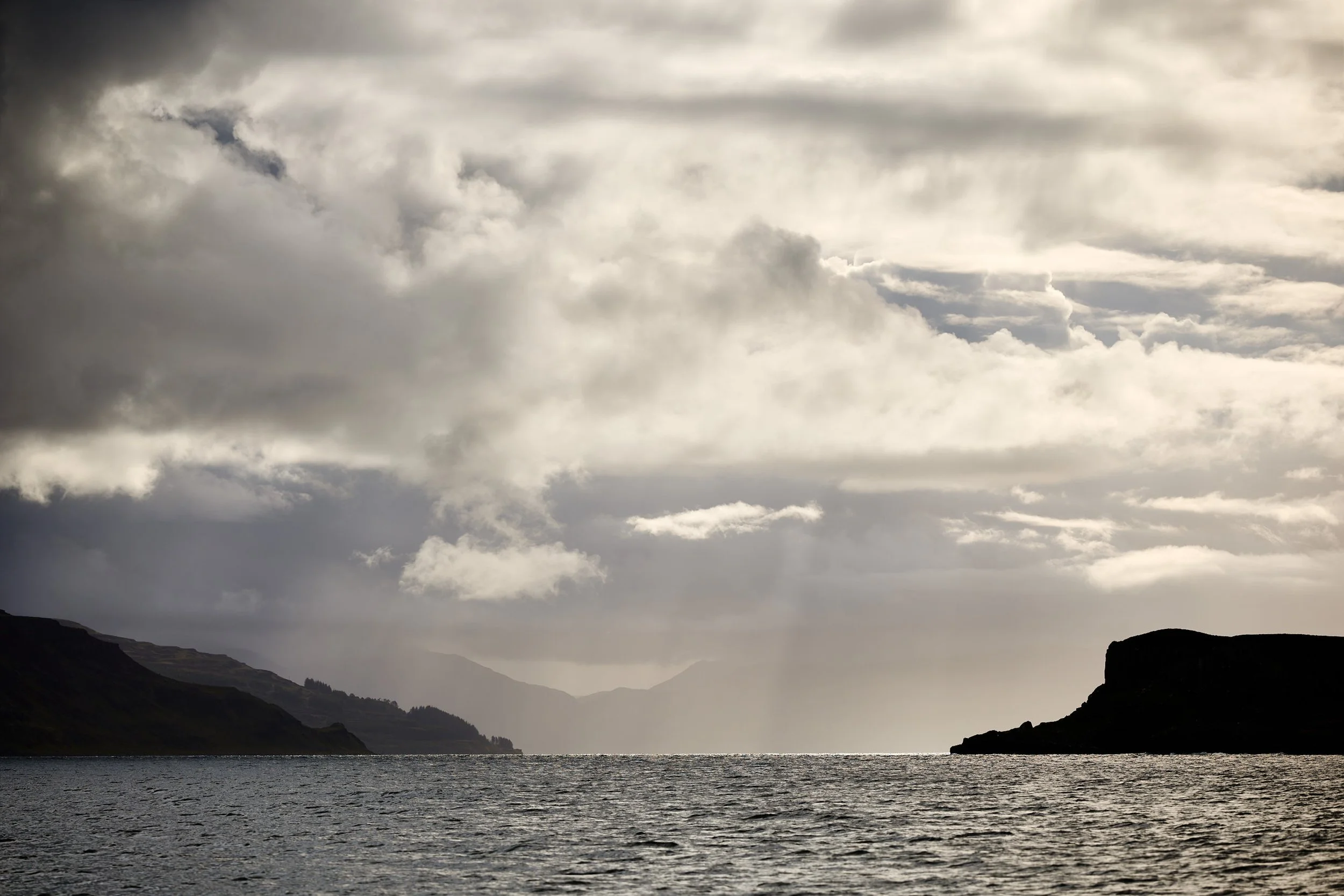 Rugged coastline with cliffs, rolling clouds, and a body of water, under a mostly cloudy sky.