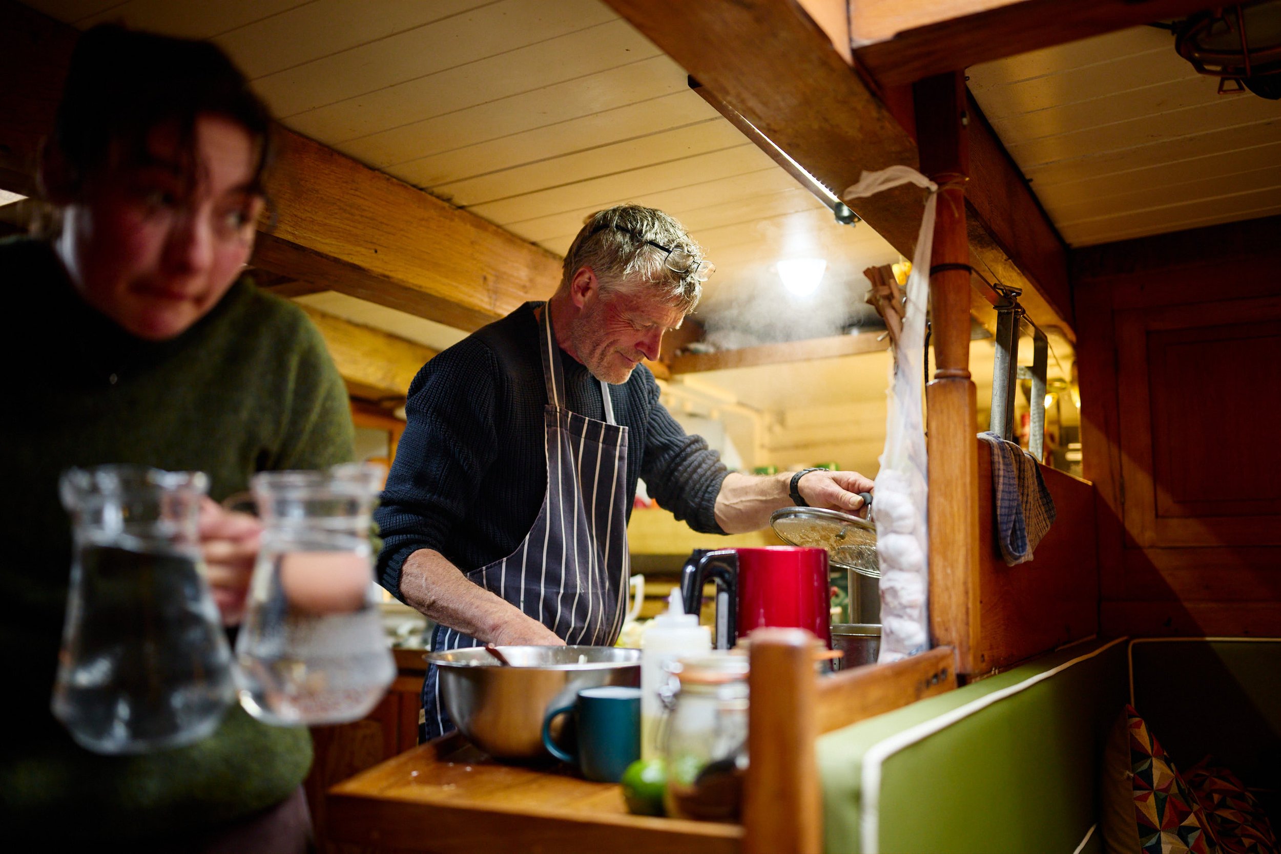 Skipper wearing glasses and an apron cooking in a cozy, wood-paneled galley with warm lighting, alongside a woman whose face is partially blurred.