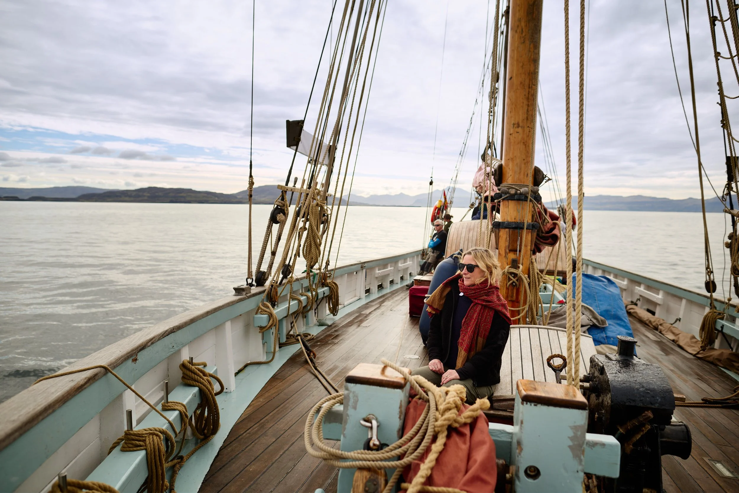 View of the deck of a sailing ship on a cloudy day with two women sitting on the deck, ropes and wooden beams visible, and a distant landmass across the water.