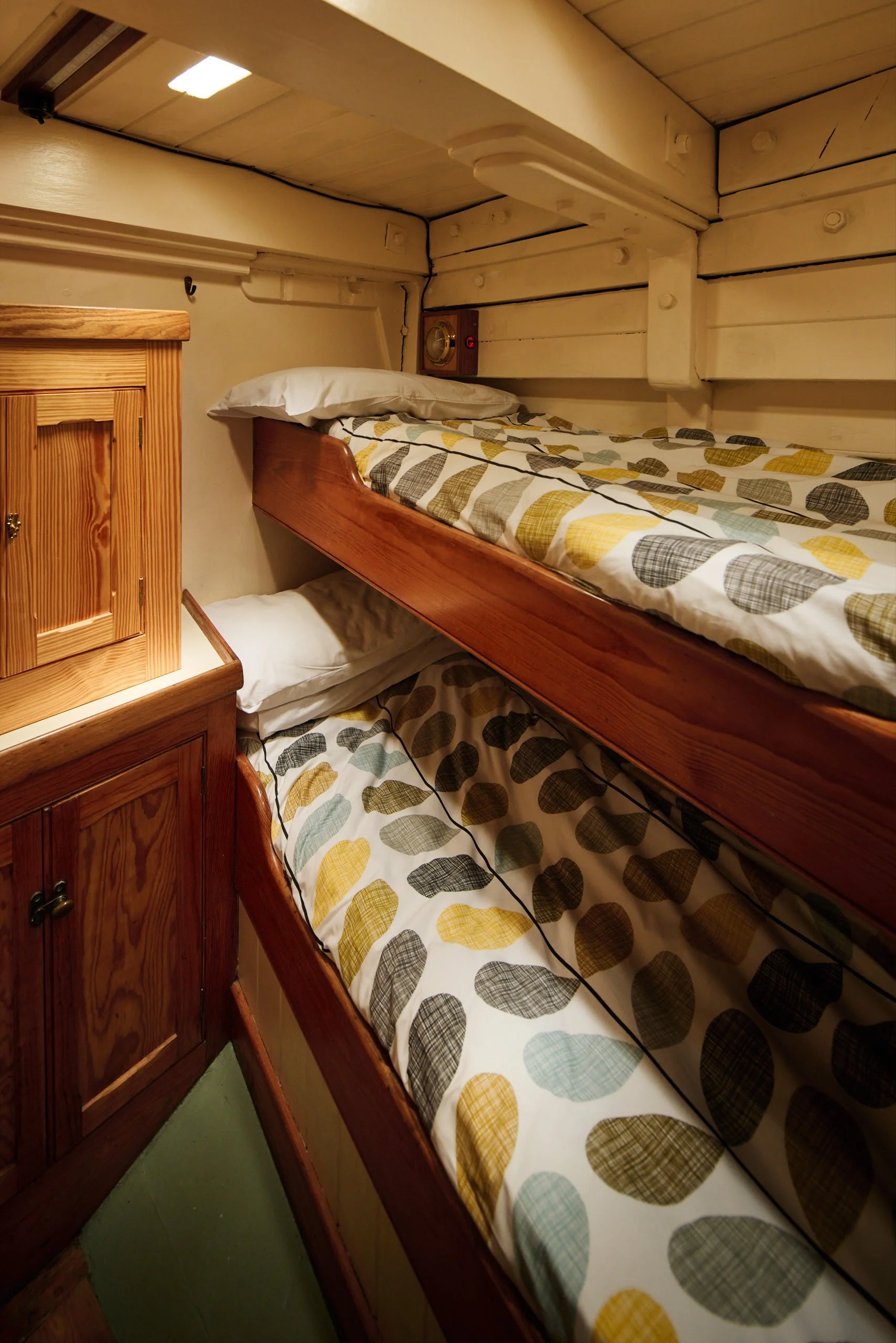 Bunk beds in a small room with wooden walls, featuring a single bed with a colorful polka dot patterned bedspread and pillows, a wooden nightstand, and an electronic clock tucked into the wall.