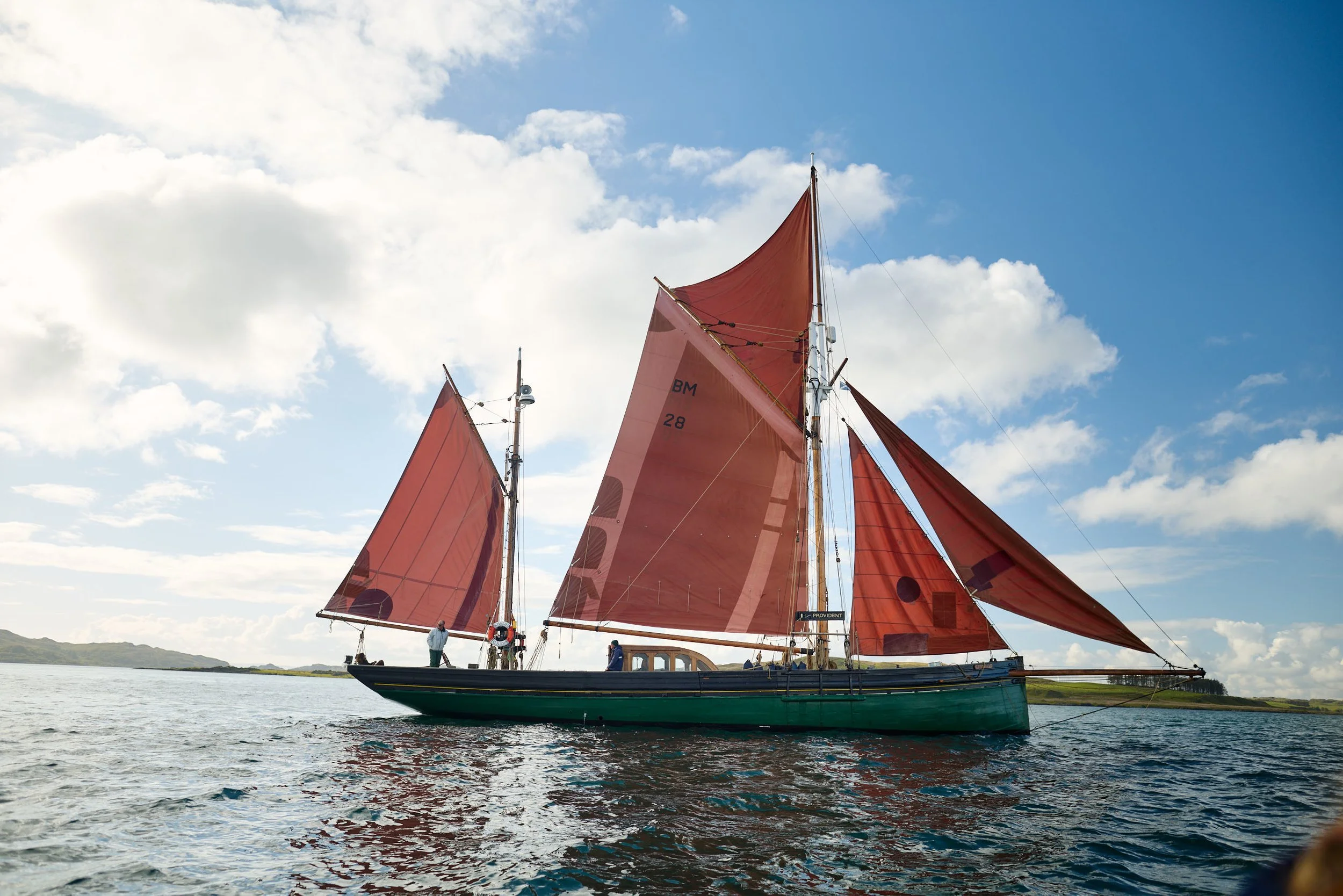 A sailboat with red sails on calm water under partly cloudy sky, with people onboard and distant land in background.