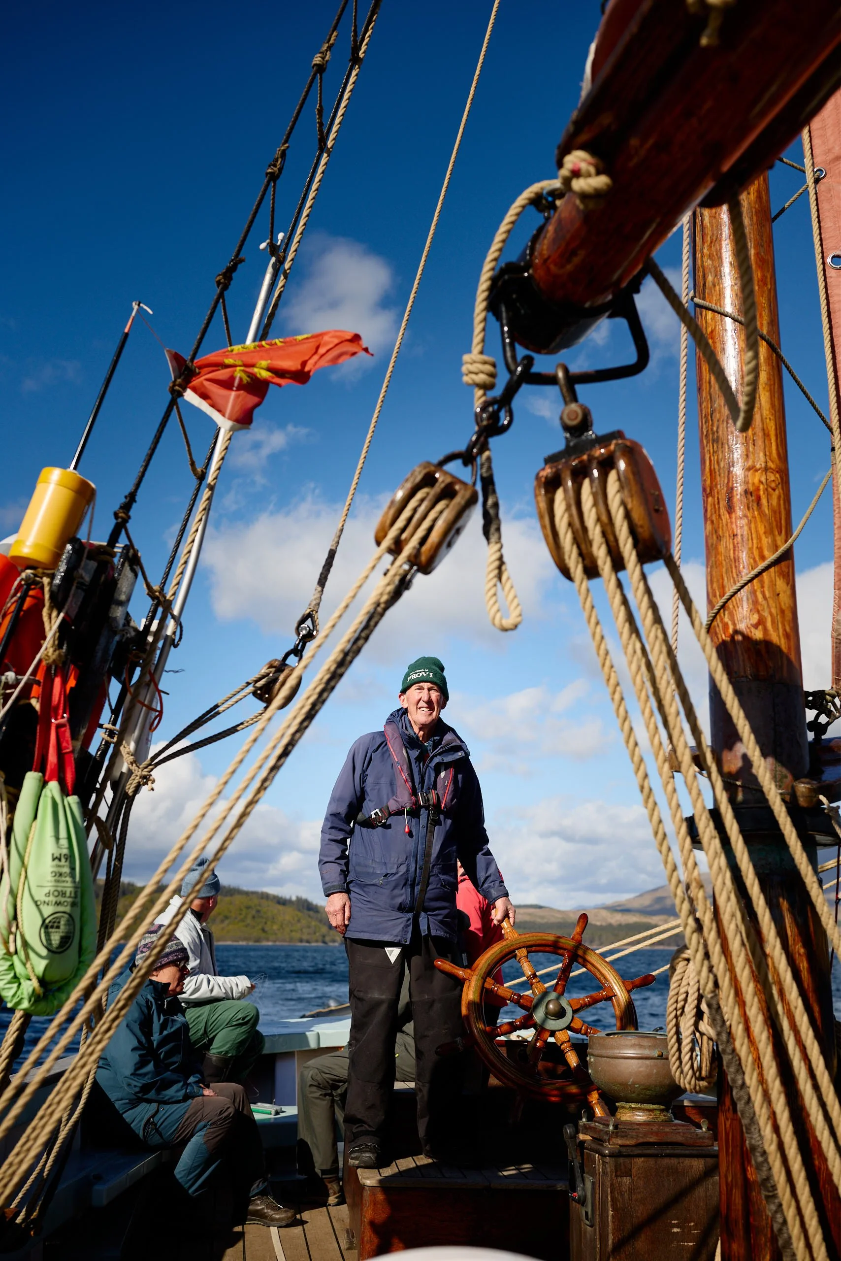 A person standing on a sailing ship's deck, holding a ship's wheel, with other crew members seated nearby. The ship has wooden railings and rigging, with a flag flying in the wind. The background shows water and distant land under a partly cloudy sky