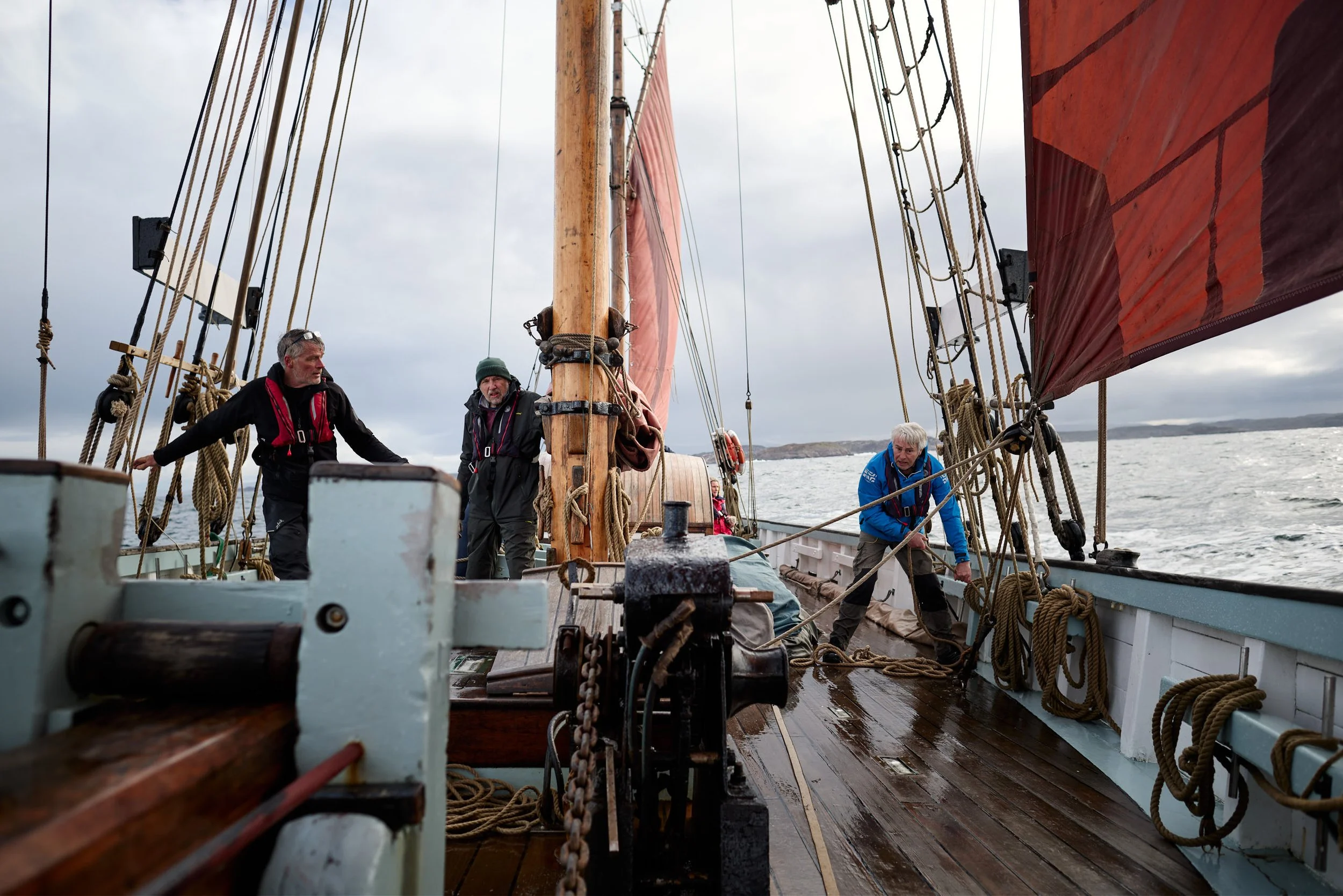 Three men are aboard a sailing ship with a wooden deck, surrounded by numerous ropes and rigging. They appear to be working or adjusting the sails, with overcast skies and a body of water with land visible in the background.