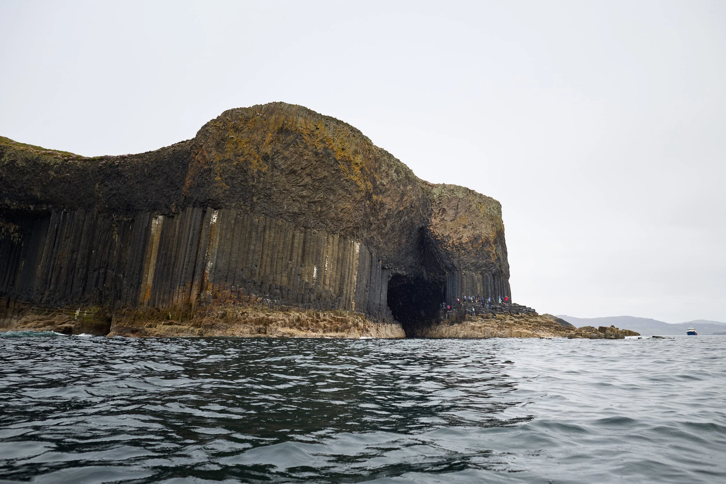 Cliffside with basalt columns and a group of people standing near the base, overlooking the ocean with a boat in the distance, overcast sky.