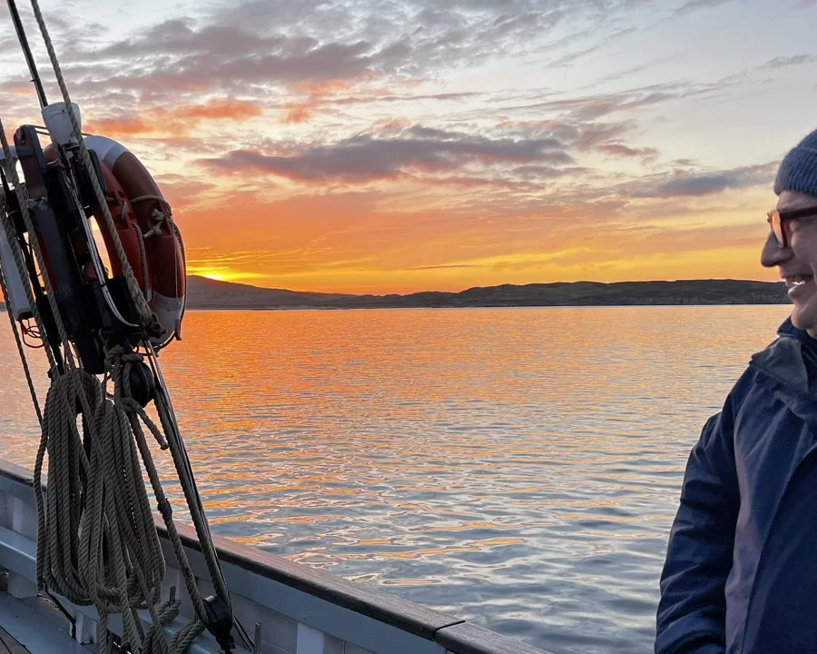 A man wearing glasses, a beanie, and a dark jacket, smiling while on a boat during sunset. The boat has safety equipment like a life preserver and ropes, with a calm body of water and a colorful sky in the background.