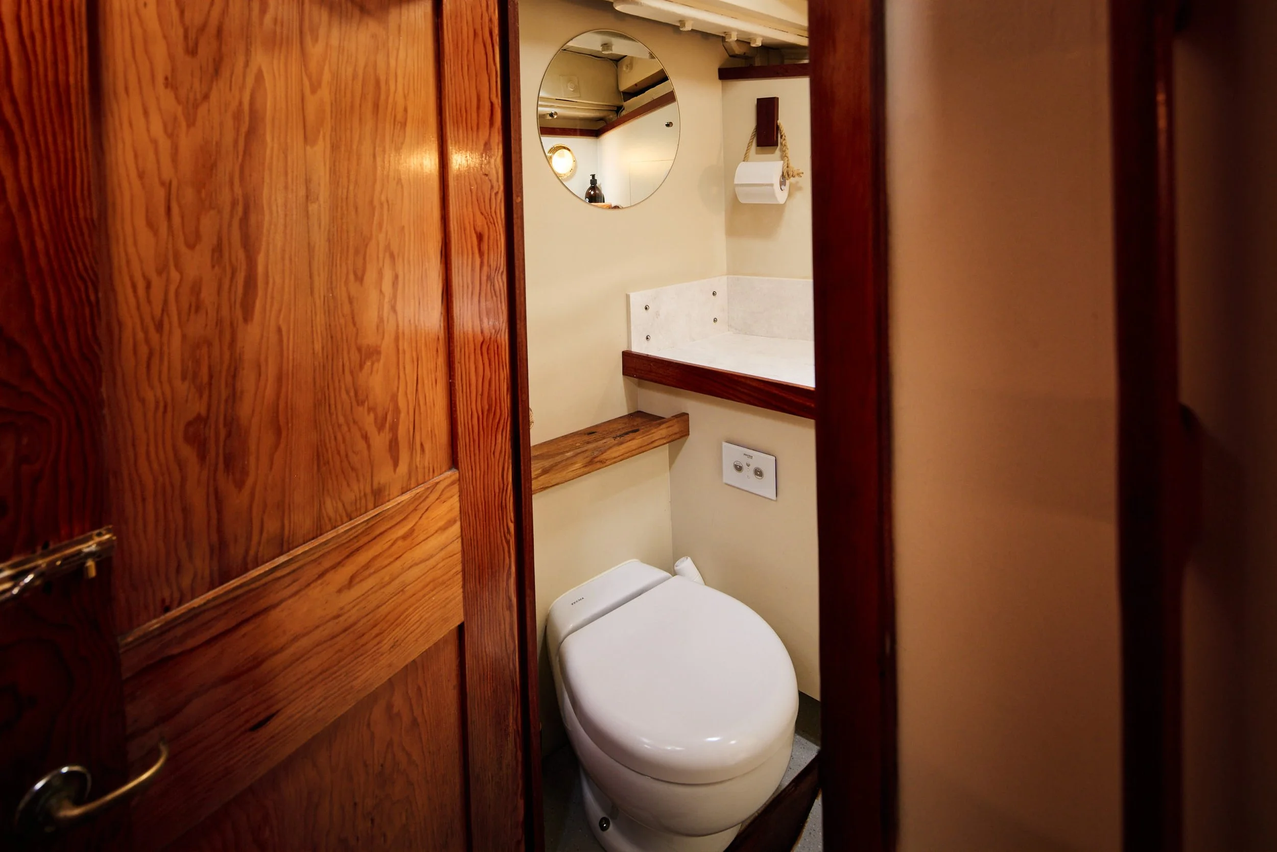 A small bathroom with a toilet, a round mirror on the wall, a toilet paper holder, and a wooden shelf. The bathroom walls are cream-colored, and there is a wooden cabinet or divider partially blocking the view.