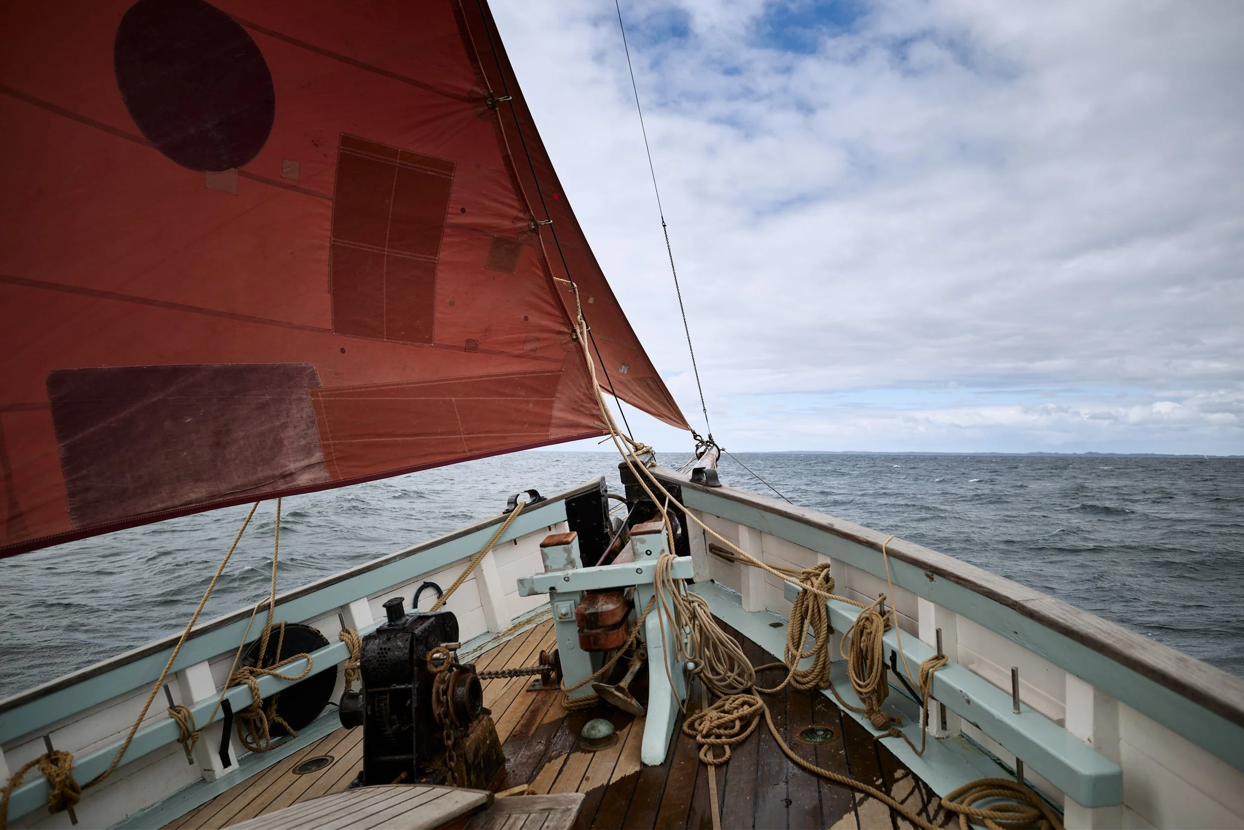 View from a boat with a large red sail on the water, ropes and equipment on the boat, and a cloudy sky in the background.