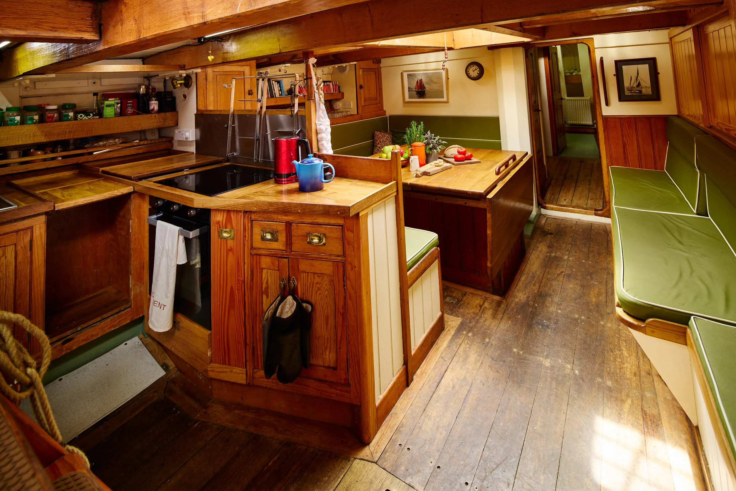 Cozy kitchen with wooden cabinets, a corner dining nook with green cushions, and a table set with apples, tomatoes, and plants, featuring wood flooring and nautical decor on the walls.