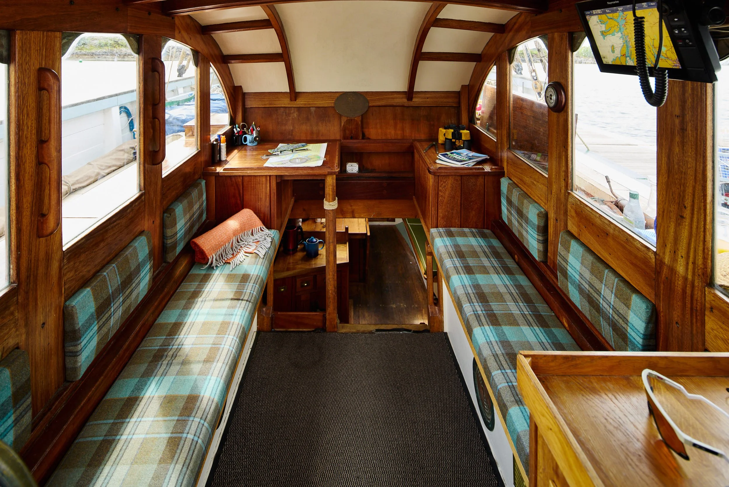 Interior of a wooden boat with plaid seating on either side, a small table, windows showing water outside, and navigation equipment.