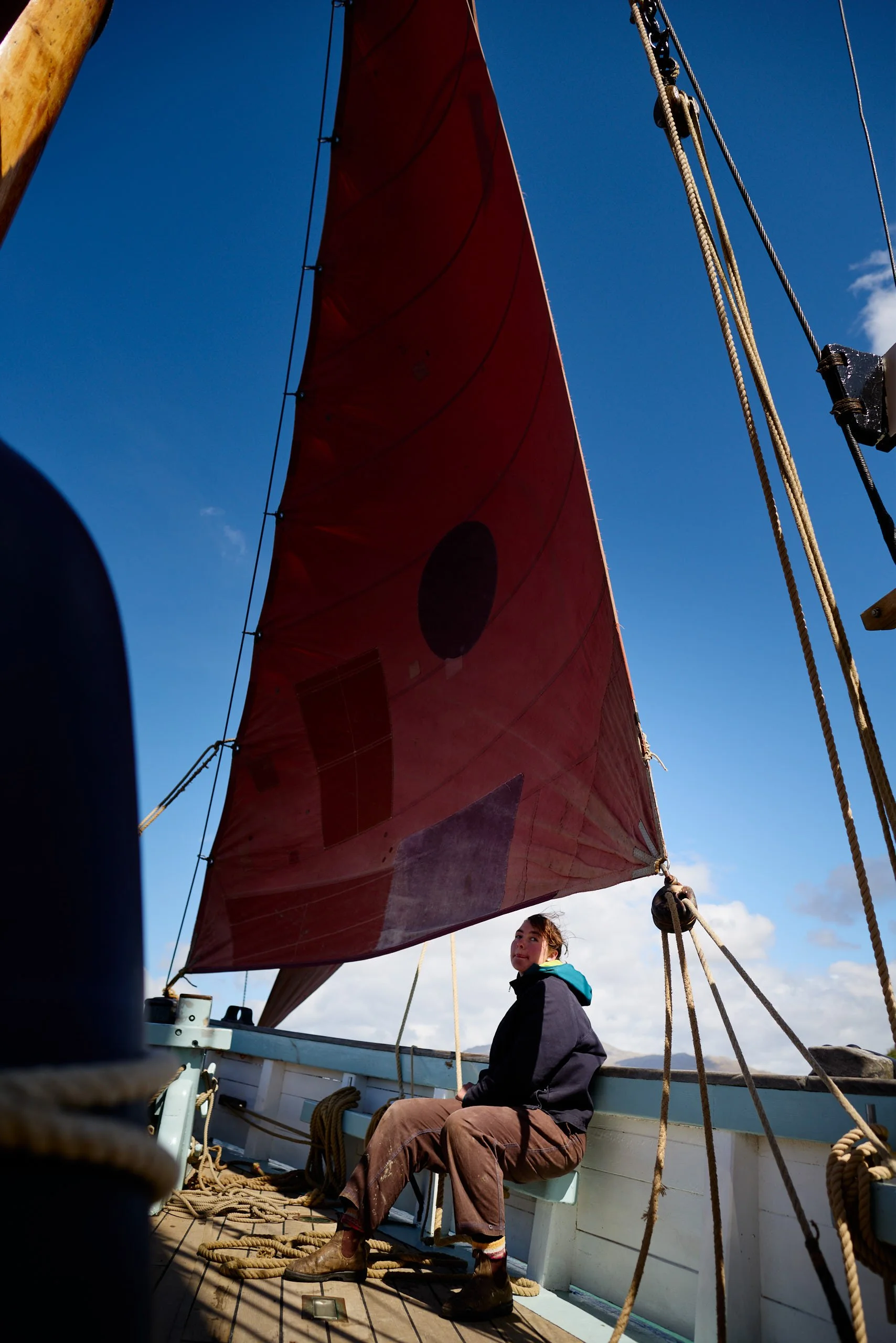 A person sitting on a boat deck beneath a large red sail with a blue sky background.