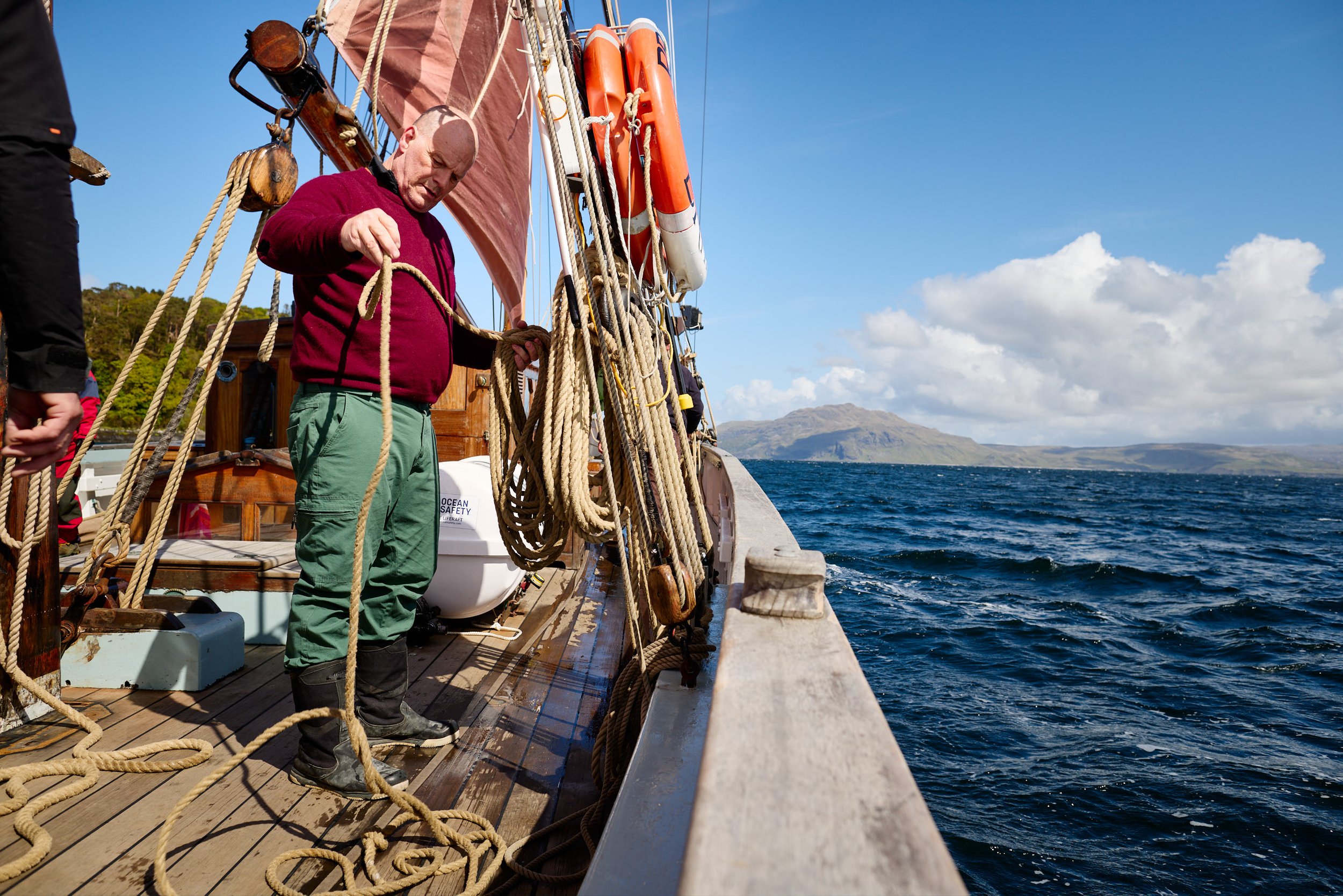A man on a sailboat is handling a thick rope on the deck, with a view of the ocean and distant mountains under a partly cloudy sky.