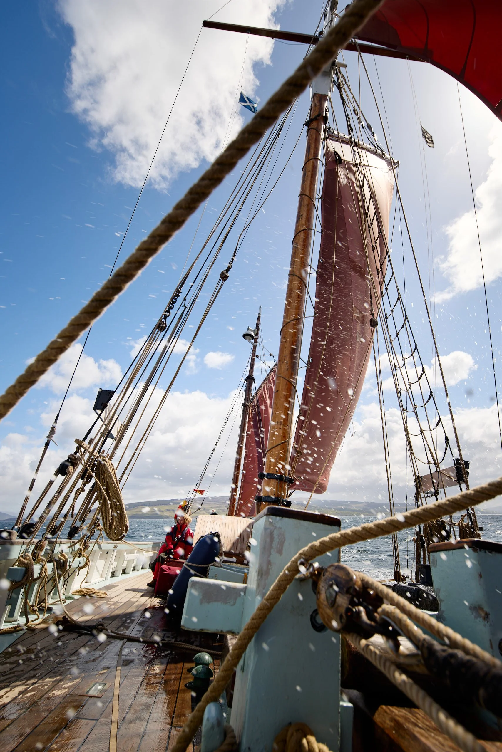 View from the deck of a sailing ship showing ropes, rigging, a mast, and the sails against a sky with scattered clouds. Part of the boat's Wooden deck and a person seated are visible at the bottom of the image. The water and distant landscape are seen below.