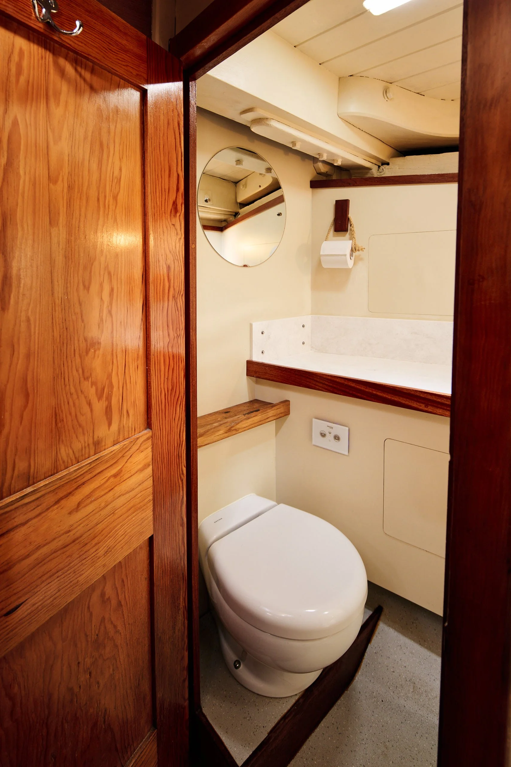 Small bathroom with a wood door partially open, showing a toilet, a small mirror, a paper towel holder with a rolled towel, a countertop, and wall outlets.