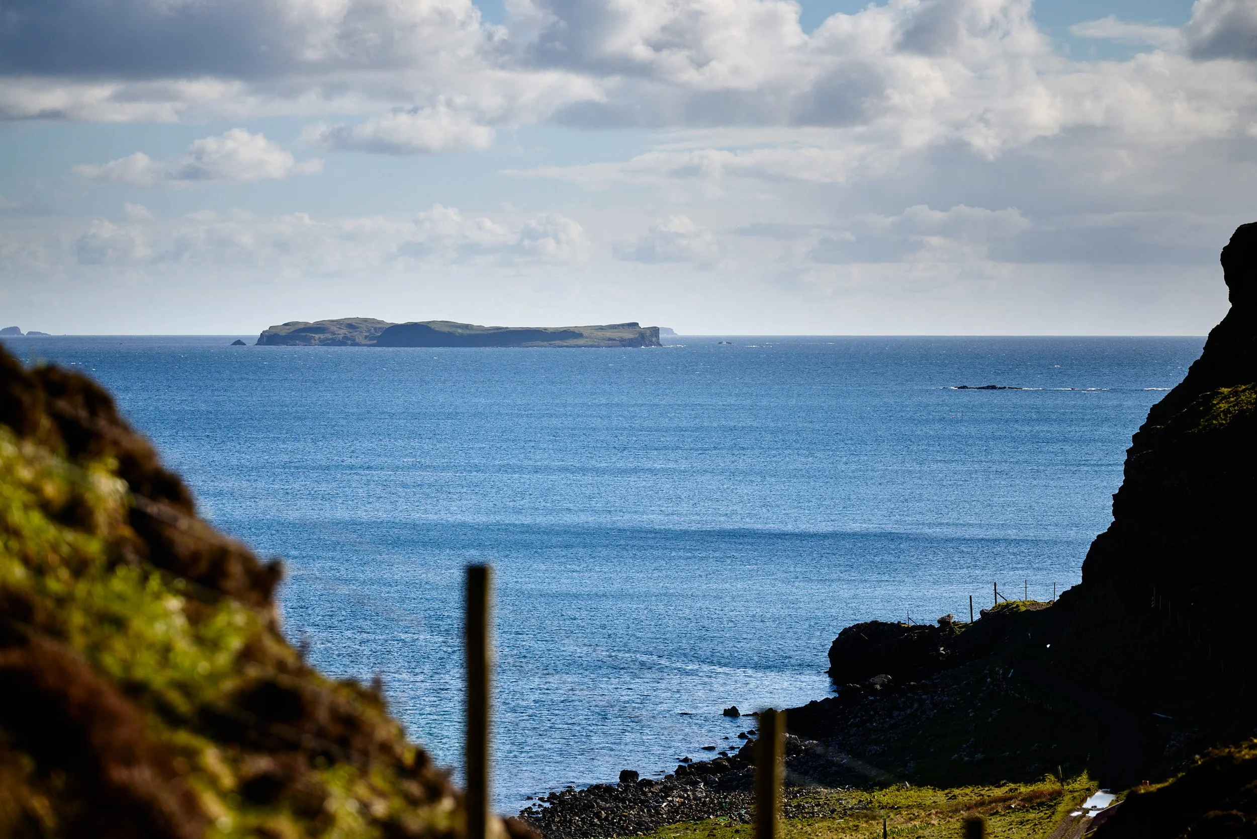 A view of the ocean with a small island in the distance, seen through a natural frame of rocky cliffs and green moss in the foreground. The sky is partly cloudy with patches of blue.