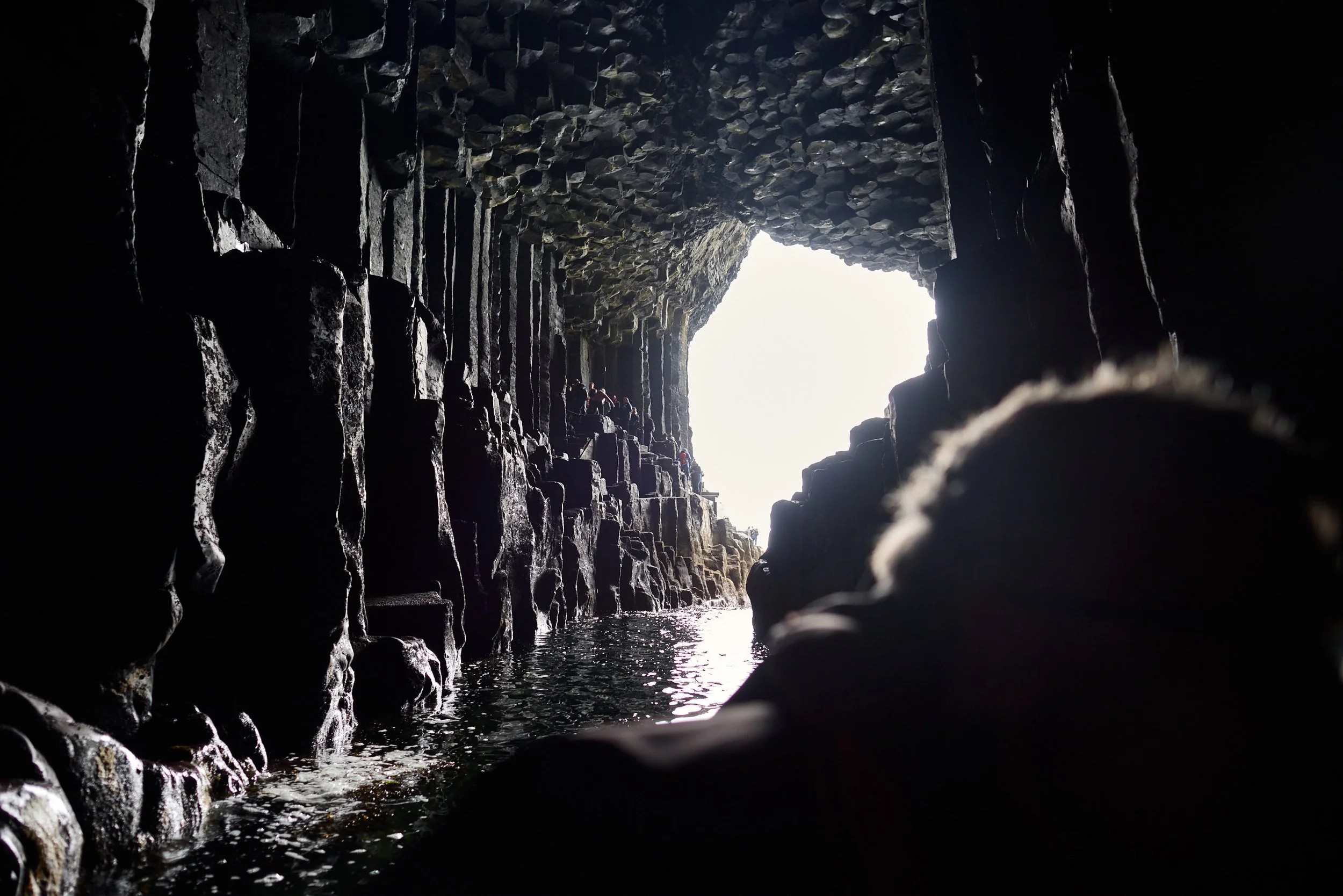 View inside Fingal's Cave with a small opening at the end, revealing some people exploring near water with rocks on the cave walls.