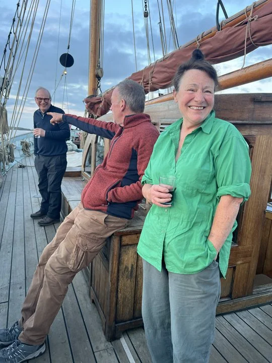 Three people on a wooden boat, two men and one woman, are enjoying a conversation. The woman is smiling and holding a glass of wine, while the man in the red jacket is leaning against the boat's railing. The man in the blue jacket is standing in the 