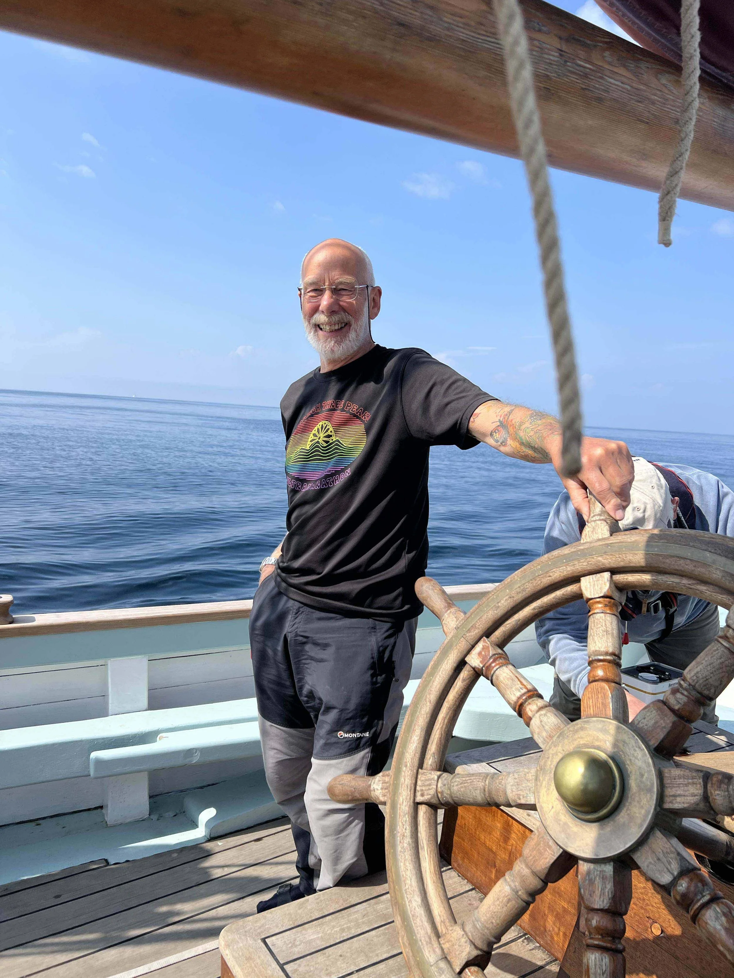 A smiling man with a beard and glasses on a boat, holding the steering wheel with the ocean and blue sky in the background.