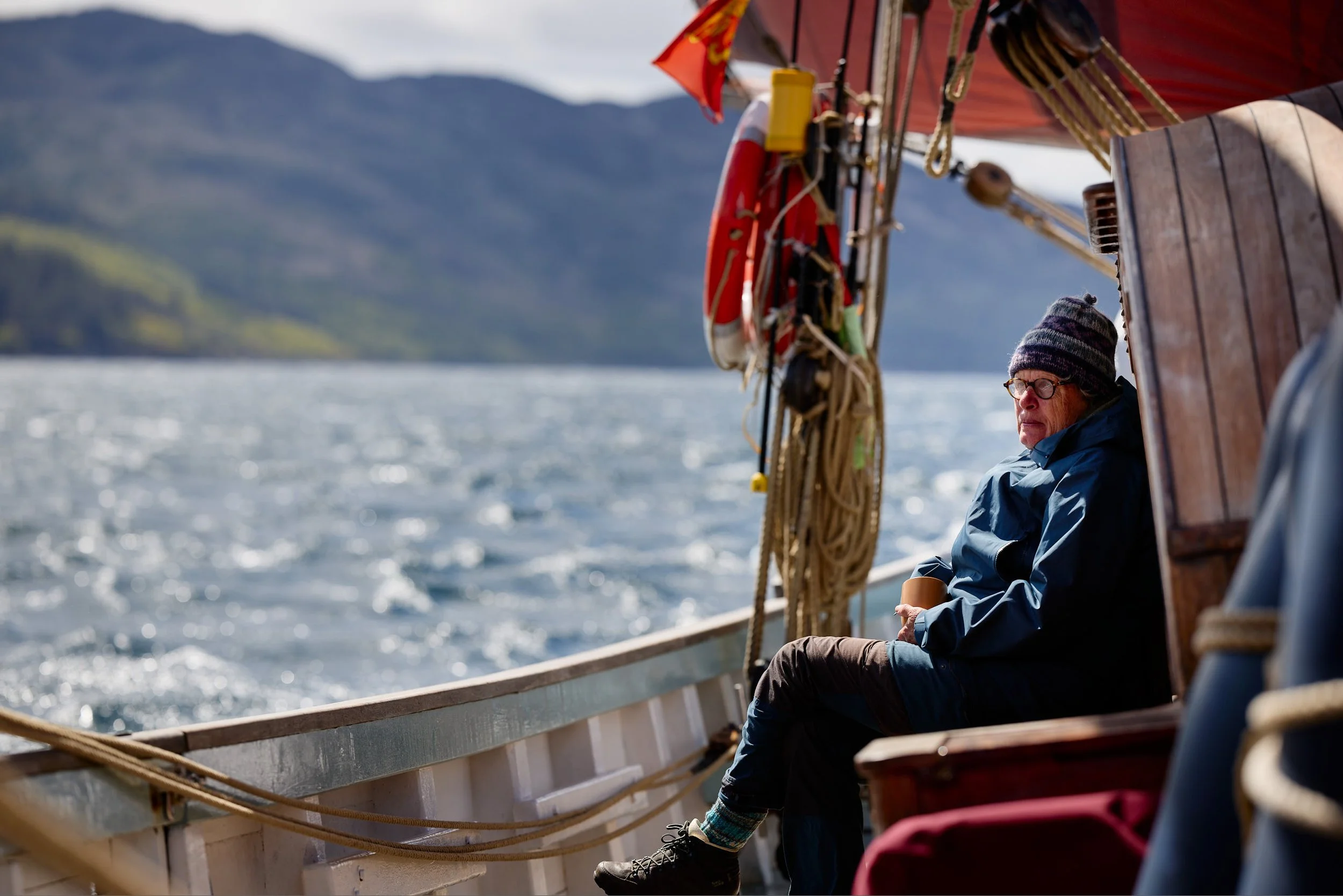 A person sitting on the edge of a boat, wearing a striped beanie, glasses, and a blue jacket, holding a cup. The boat has a red canopy, and nearby are ropes, pulleys, and a lifebuoy. Water and distant mountains are visible in the background.