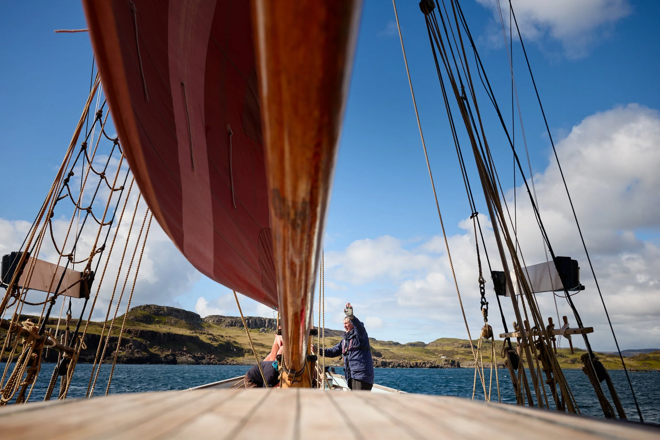 Looking up from the deck of a sailing ship with multiple ropes and rigging against a blue sky, people standing and enjoying the outdoors near the water with a hilly landscape in the background.