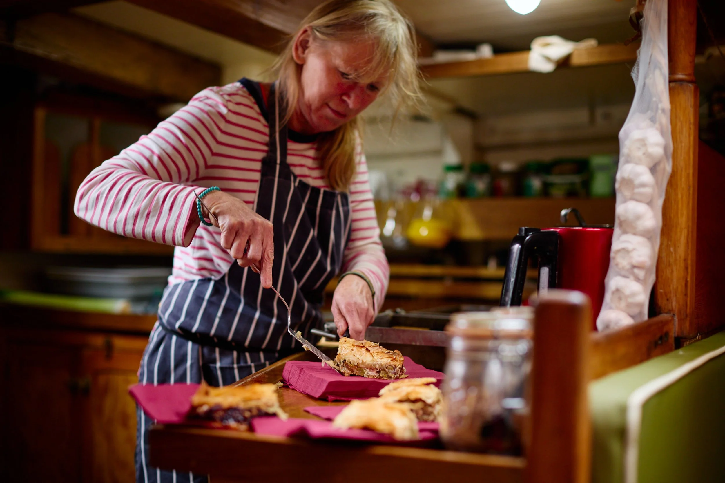 Woman with blonde hair in a striped shirt and apron slicing a piece of pie on a pink cutting board in a cozy kitchen.