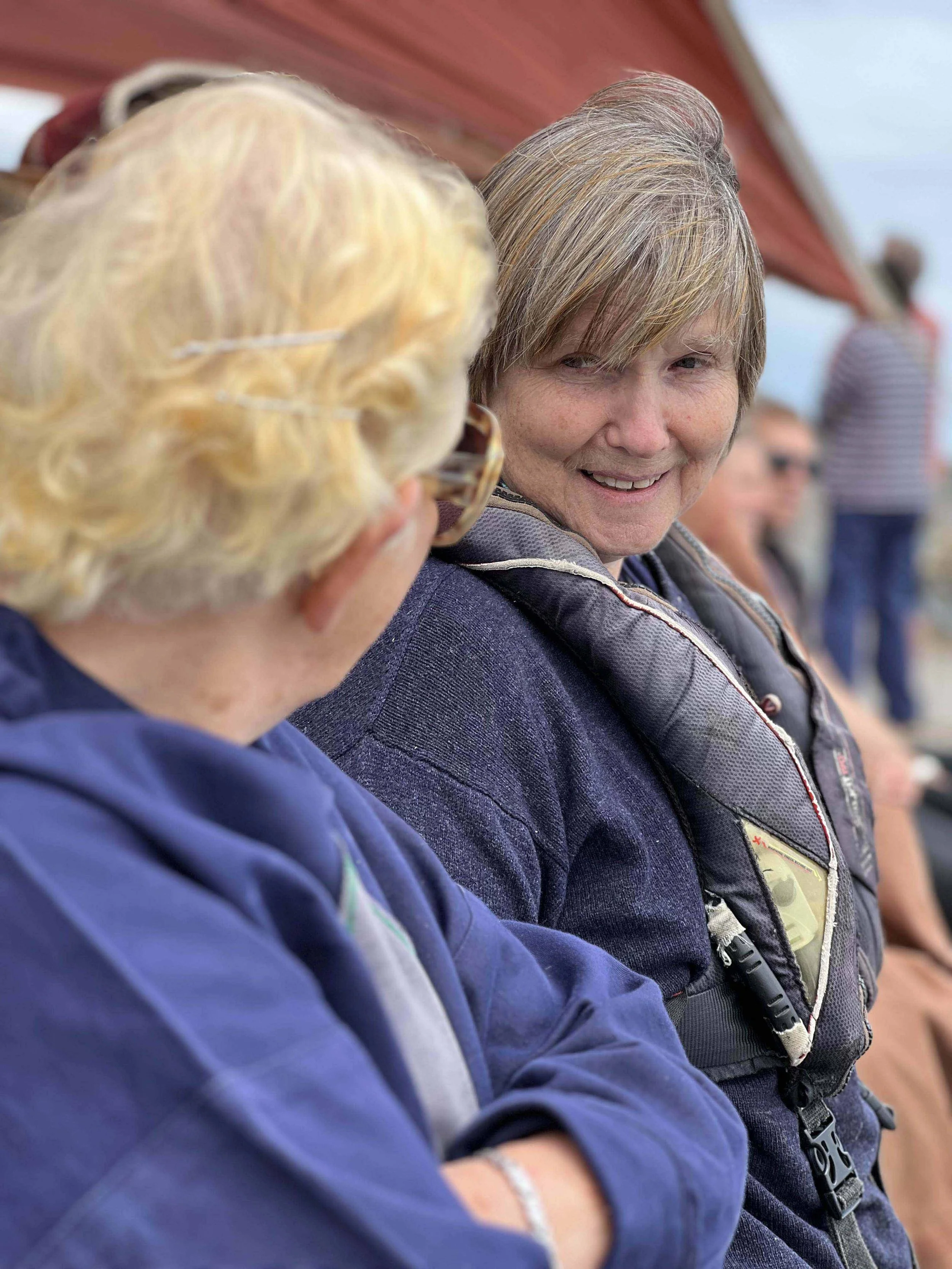 Close-up of two women sitting outdoors, smiling and engaging in conversation, with other people blurred in the background.
