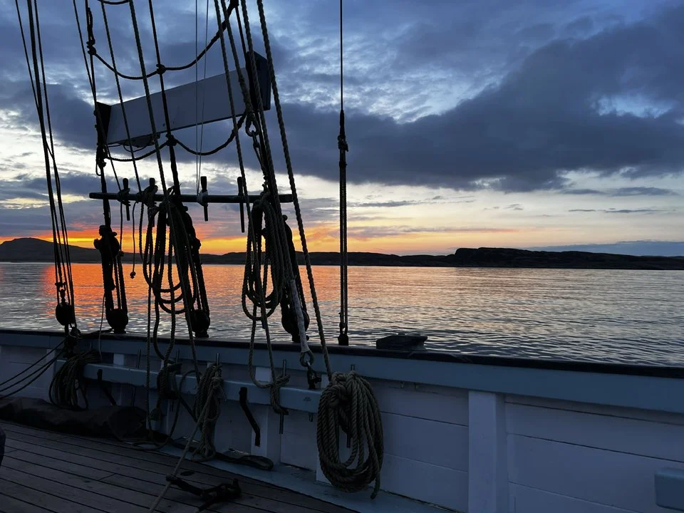 View from a Provident  at sunset with ropes and rigging in the foreground, calm water, and a distant shoreline under a partly cloudy sky on the Scottish Islands