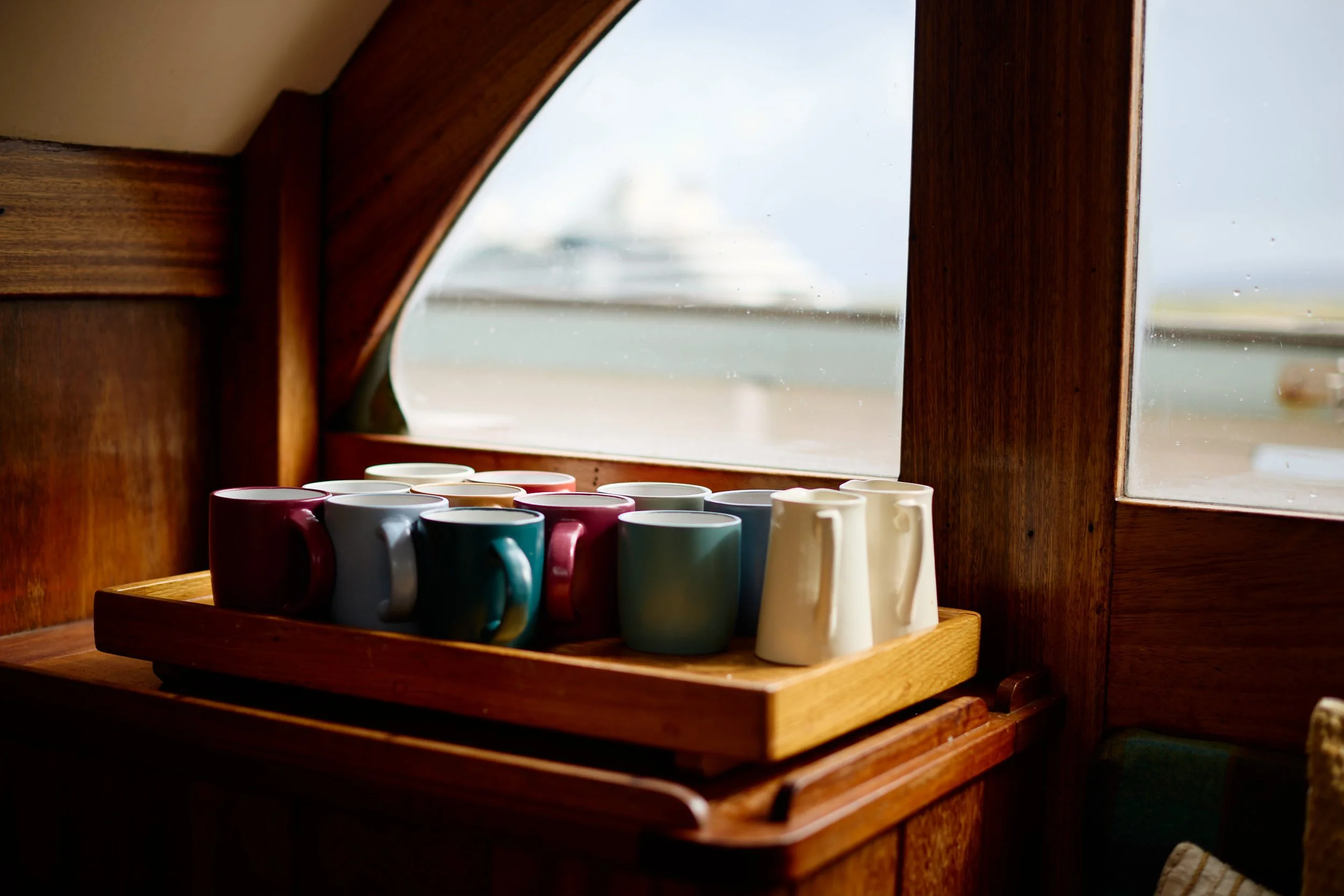 Colorful ceramic mugs arranged on a wooden tray on a windowsill in a cozy wooden interior, with a view of the beach and water outside.