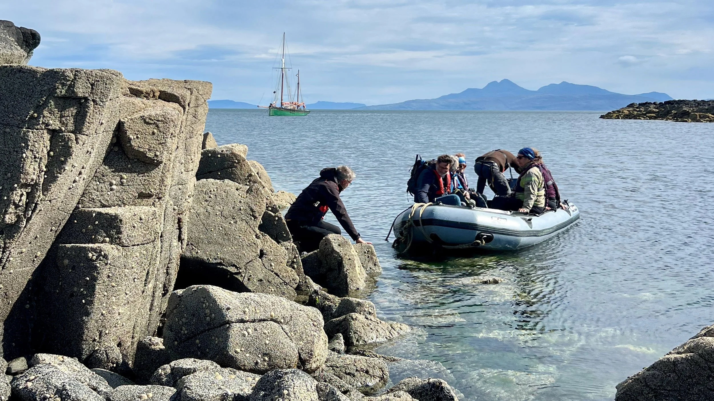 Group of people using a small inflatable boat near rocky shoreline with sailboat and mountains in background
