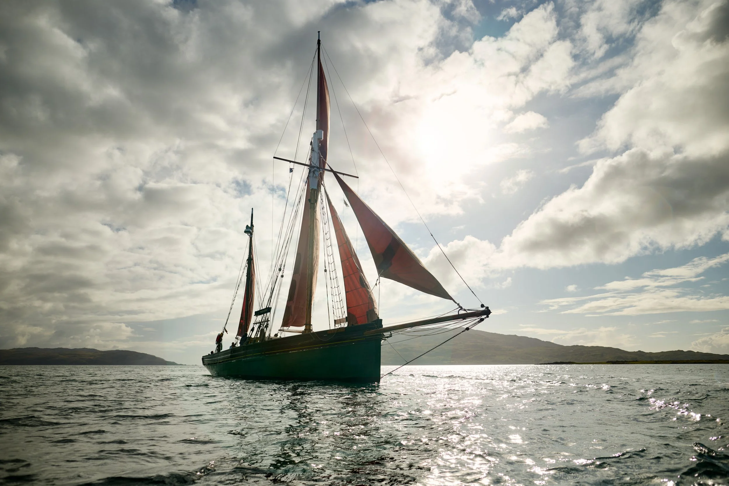 Provident with red sails floating on water under a cloudy sky with the sun shining through.
