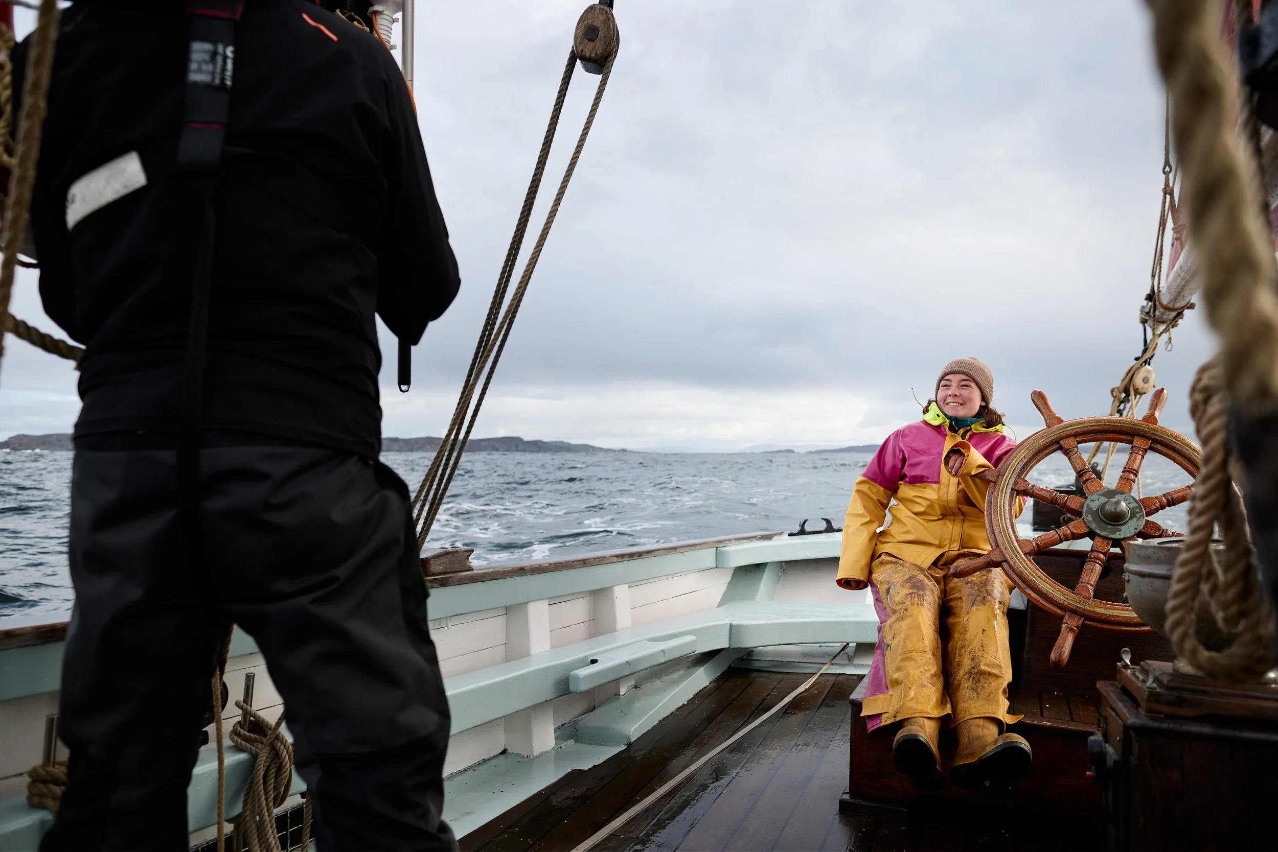 A young woman dressed in yellow rain gear and a beanie sitting at the ship's steering wheel, smiling. A person in dark clothing stands nearby on a boat, with ocean and cloudy sky in the background.